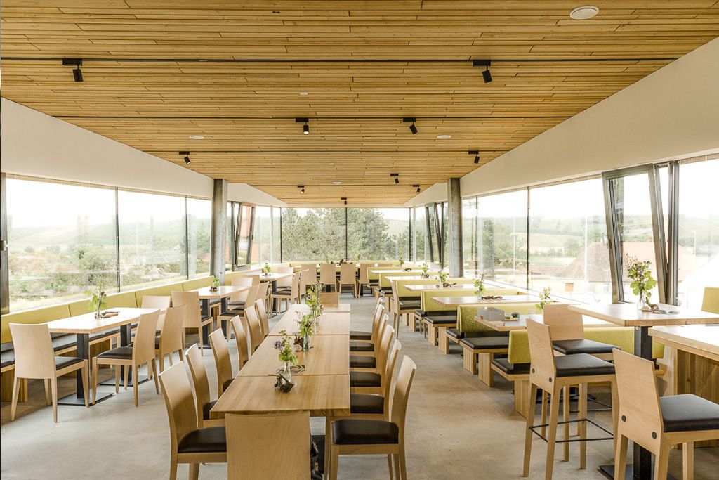 Pohled do interiéru moderní restaurace s dřevěným nábytkem a velkými okny.