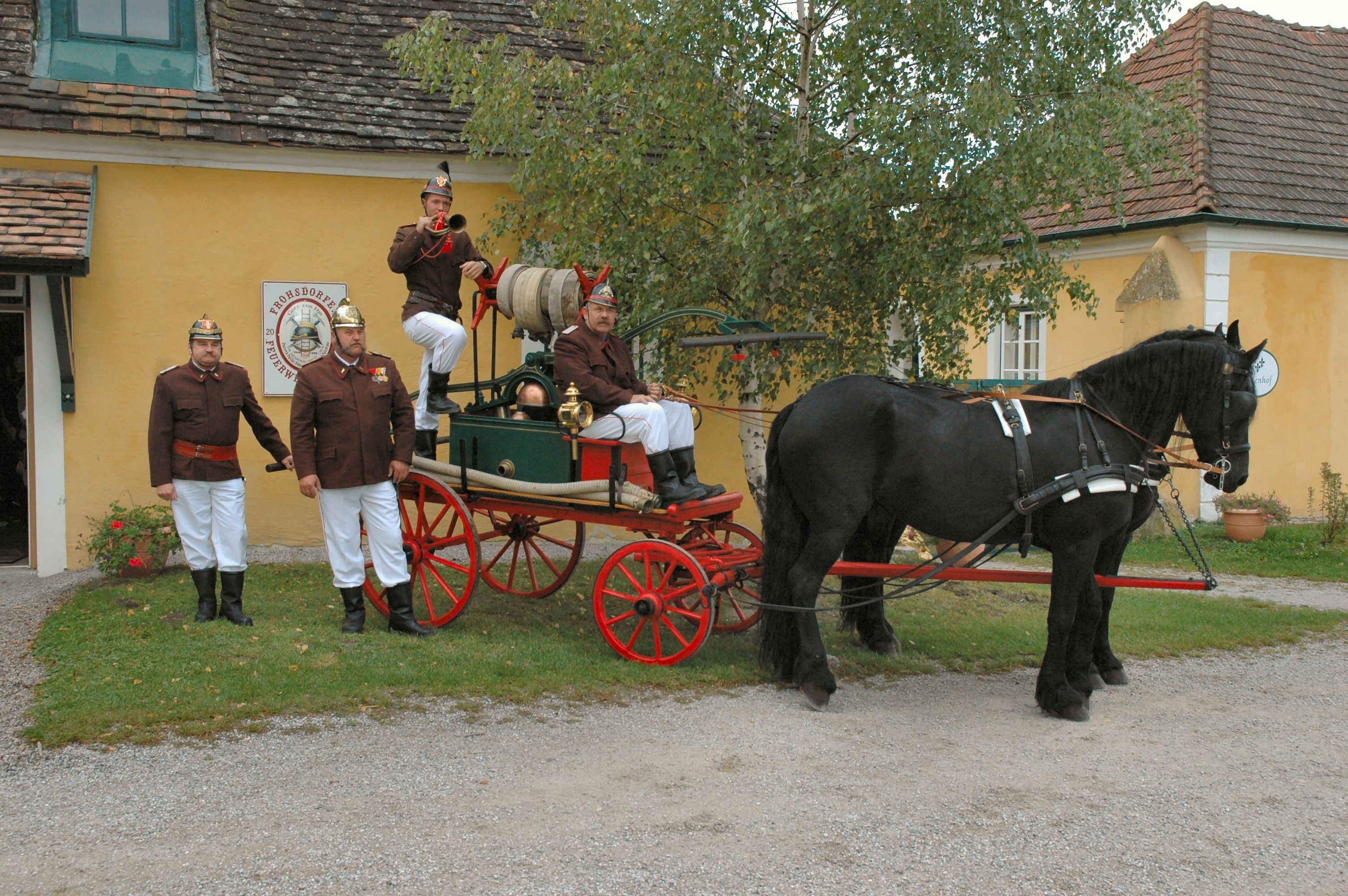 Historické hasičské auto s koněm a hasiči v uniformách před žlutou budovou.
