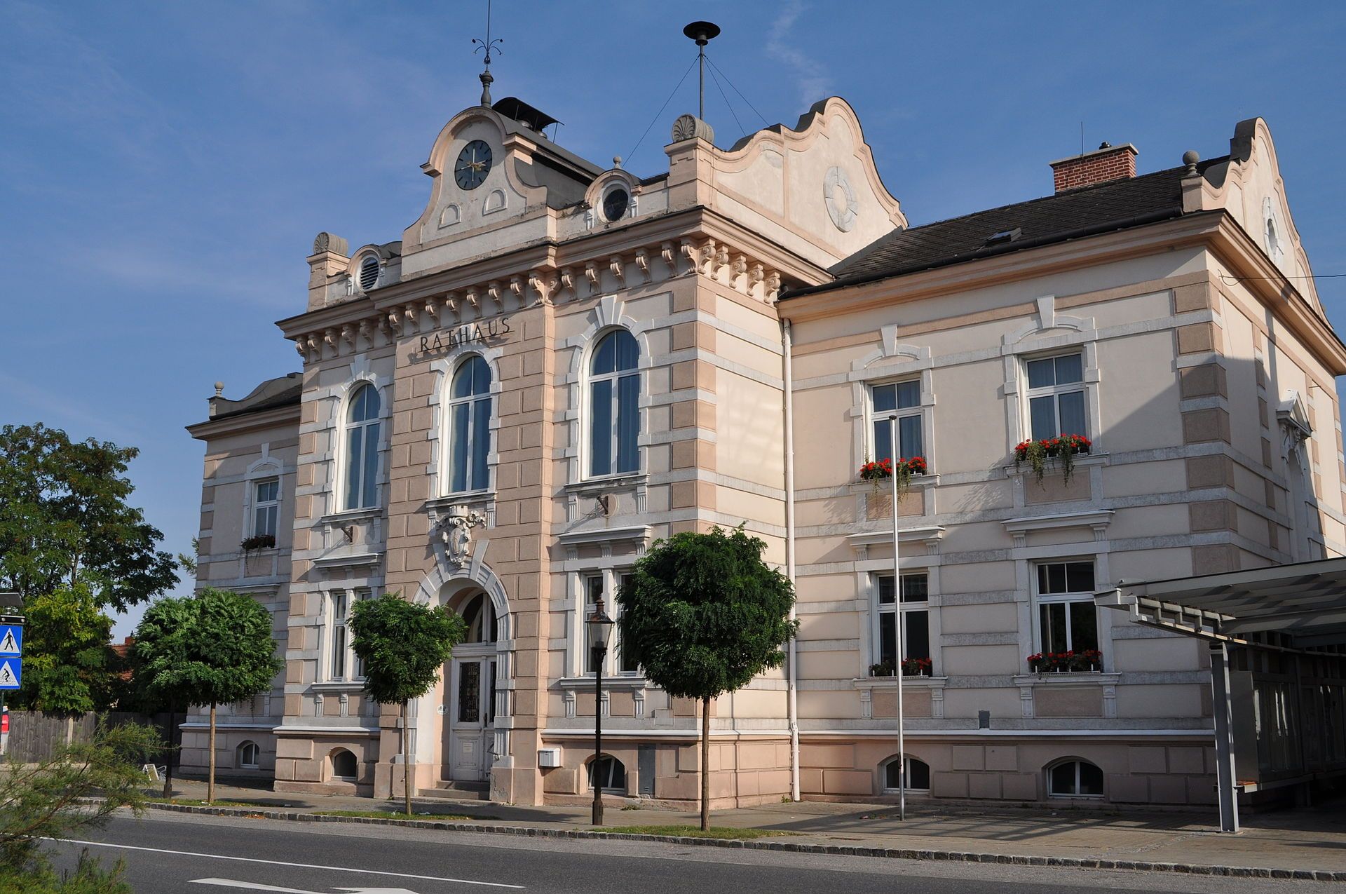 Felixdorfská radnice, historická budova s hodinovou věží a zdobenými okny, obklopená stromy a ulicí v popředí.