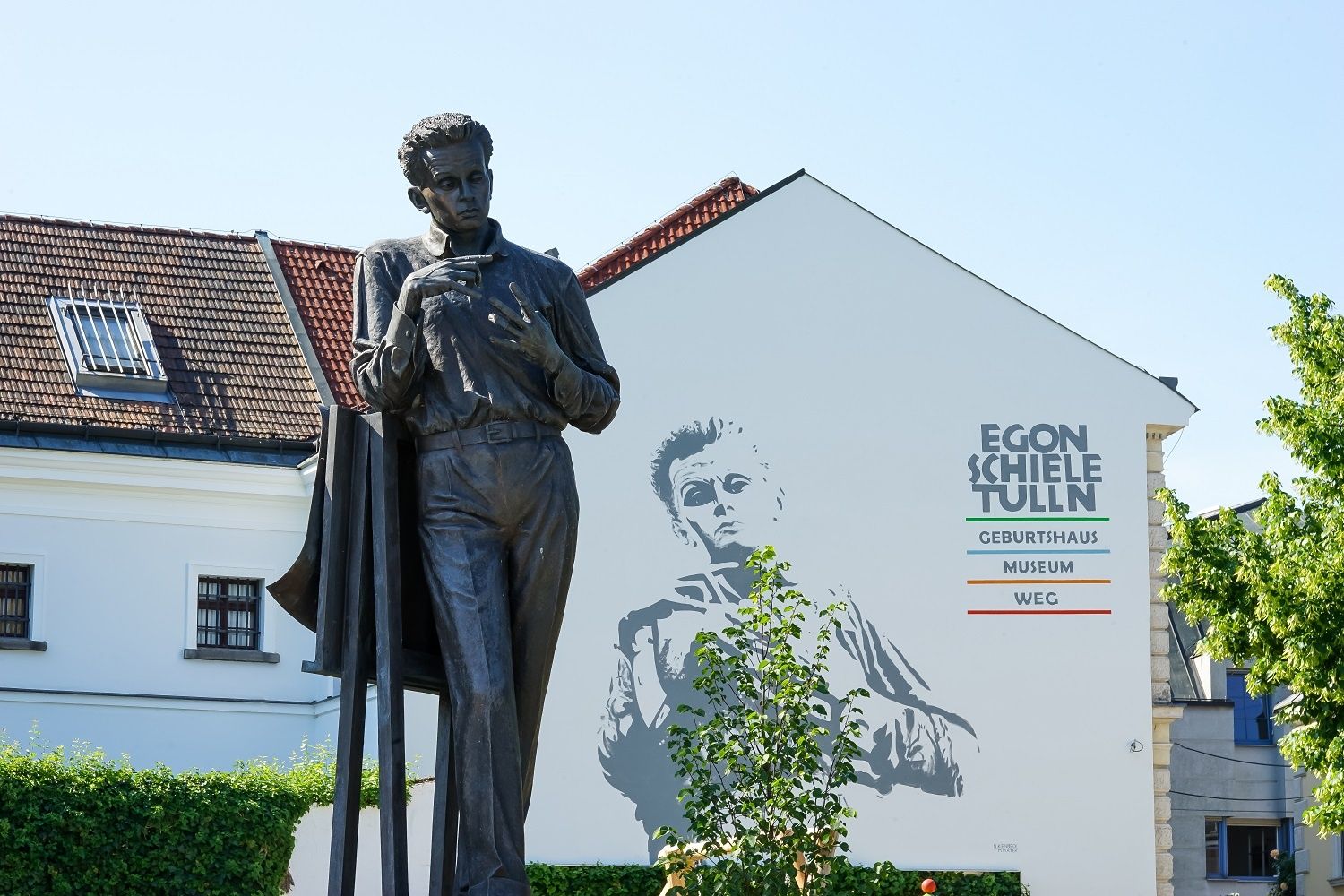 Socha Egona Schieleho před budovou s nástěnnou malbou a nápisem "Egon Schiele Tulln".