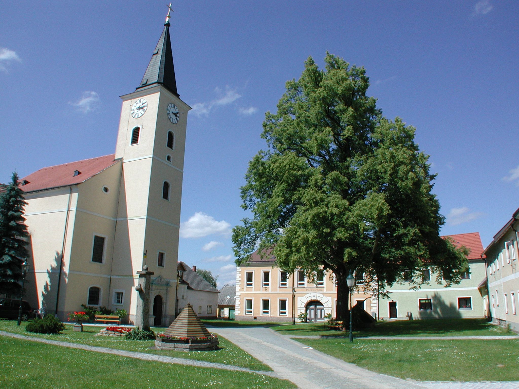 Kostel s věží a hodinami na návsi se stromy a budovami.