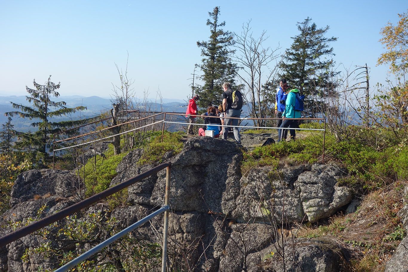 Skupina turistů na skalní vyhlídce se zábradlím, obklopená stromy a horami v pozadí.