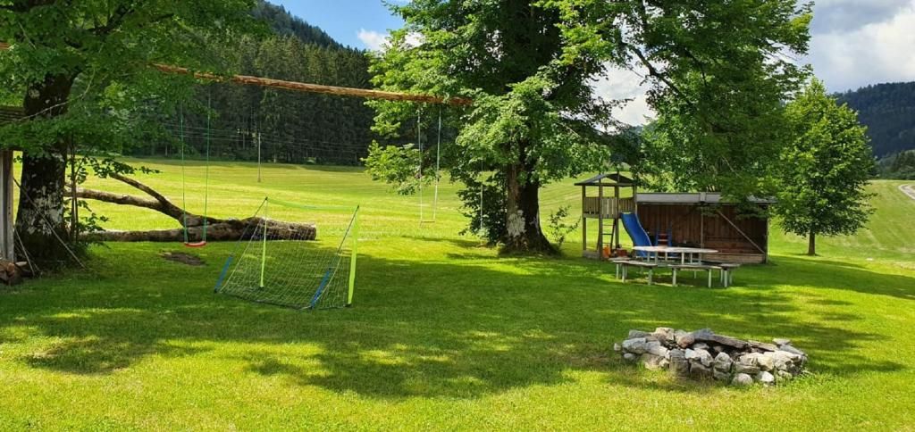 Dětské hřiště na zelené louce s houpačkami, prolézačkou, skluzavkou a ohništěm. V pozadí jsou vidět stromy a kopce.