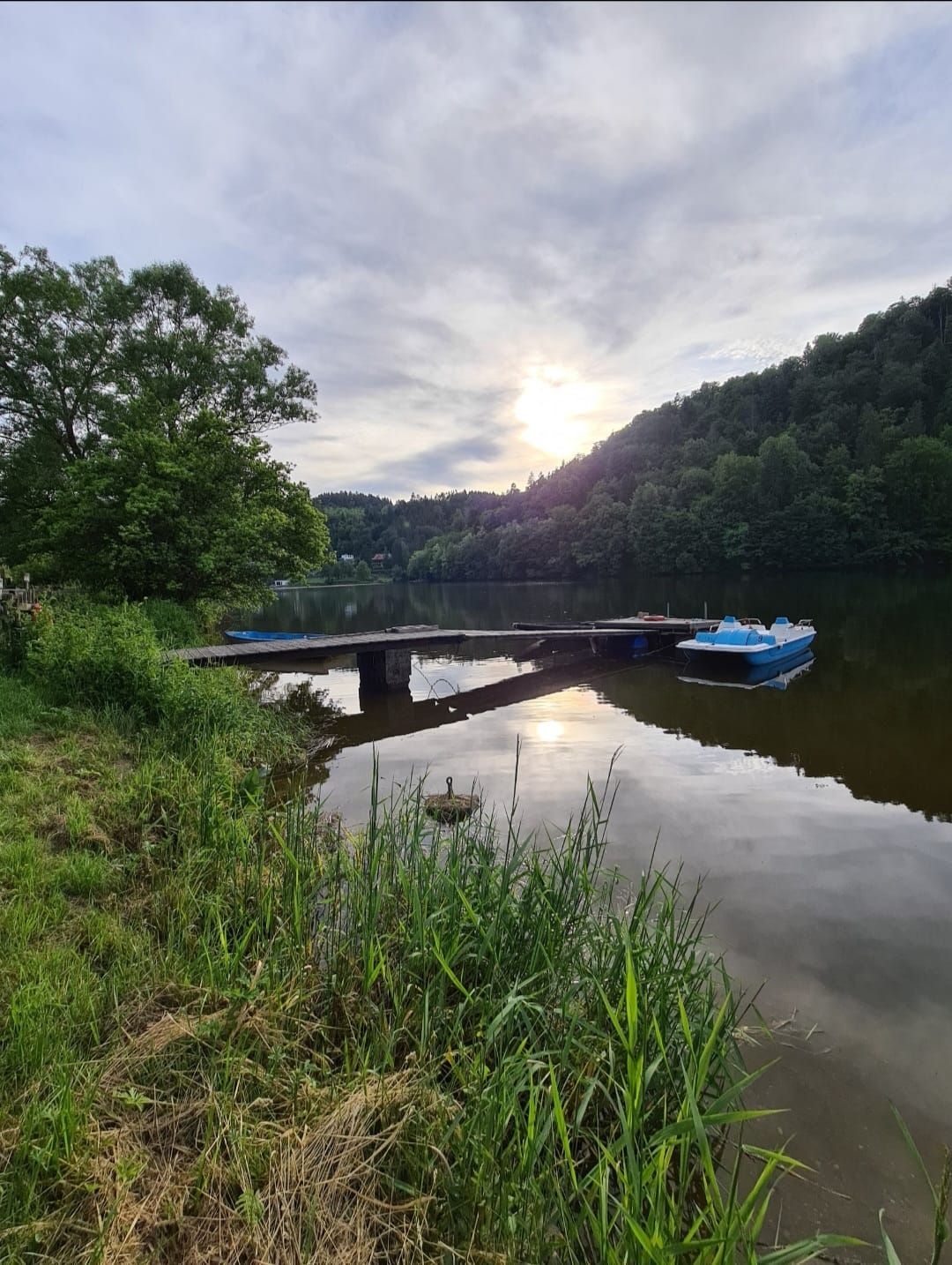 Klidná nádrž s molem a šlapadlem ve vodě, obklopená zelenými stromy a zataženou oblohou.