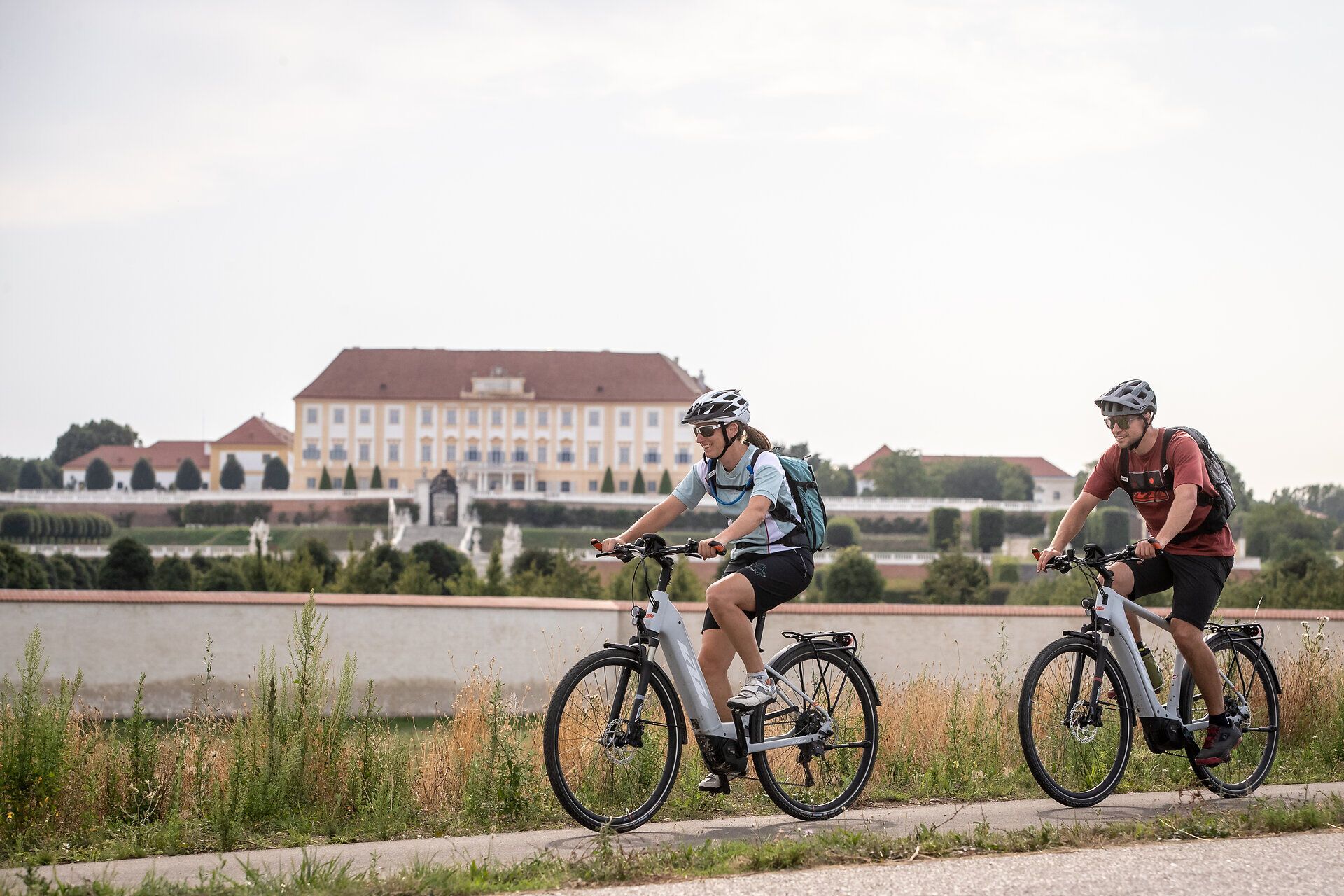 Dva cyklisté si užívají čerstvého vzduchu a malebné krajiny podél klidného kanálu. Kopce v pozadí a bujná vegetace vytvářejí příjemnou atmosféru pro milovníky outdoorových aktivit. Zde lze prožít léto v jeho nejkrásnější podobě.