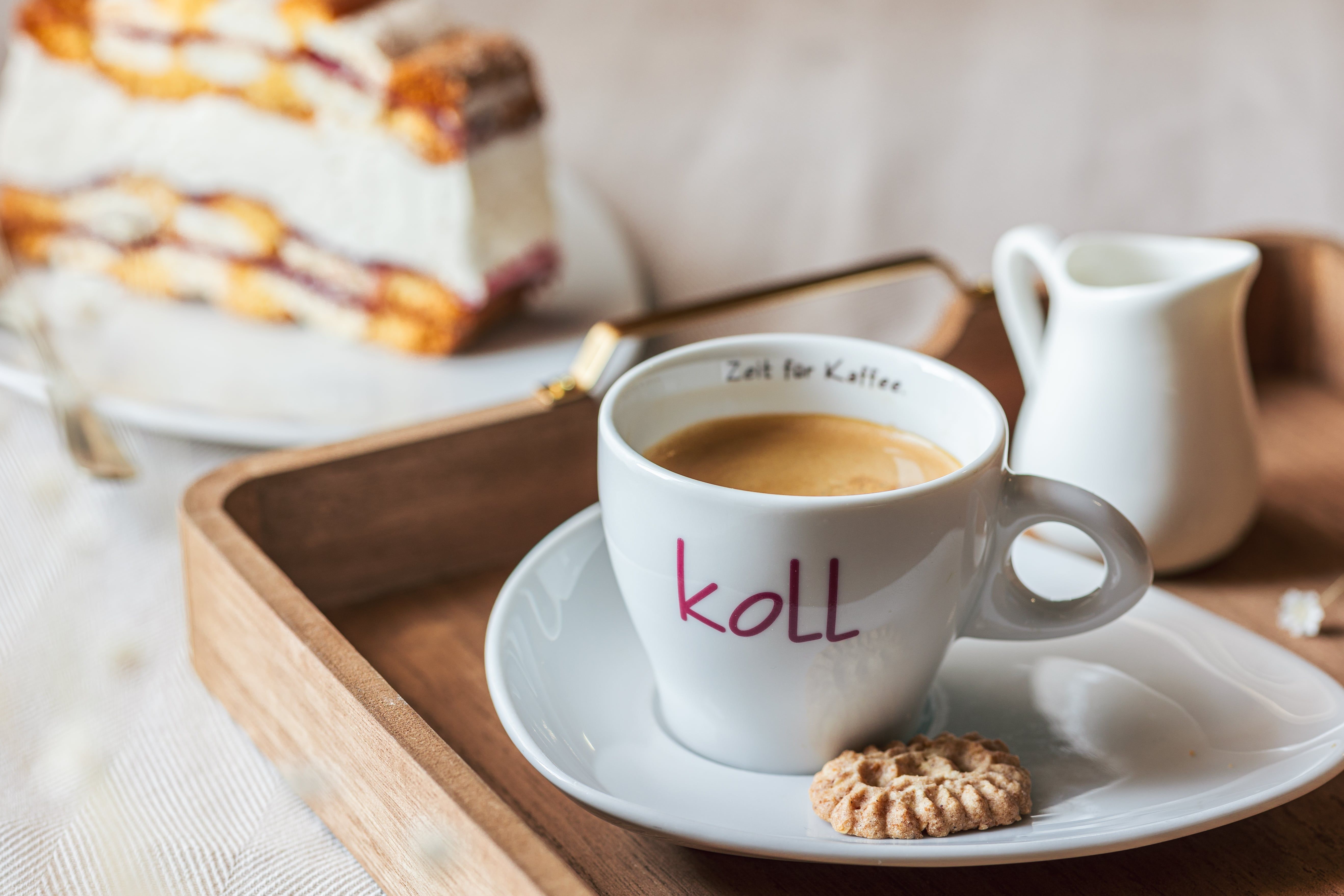 Šálek kávy se sušenkou na podnose, v pozadí kousek dortu.