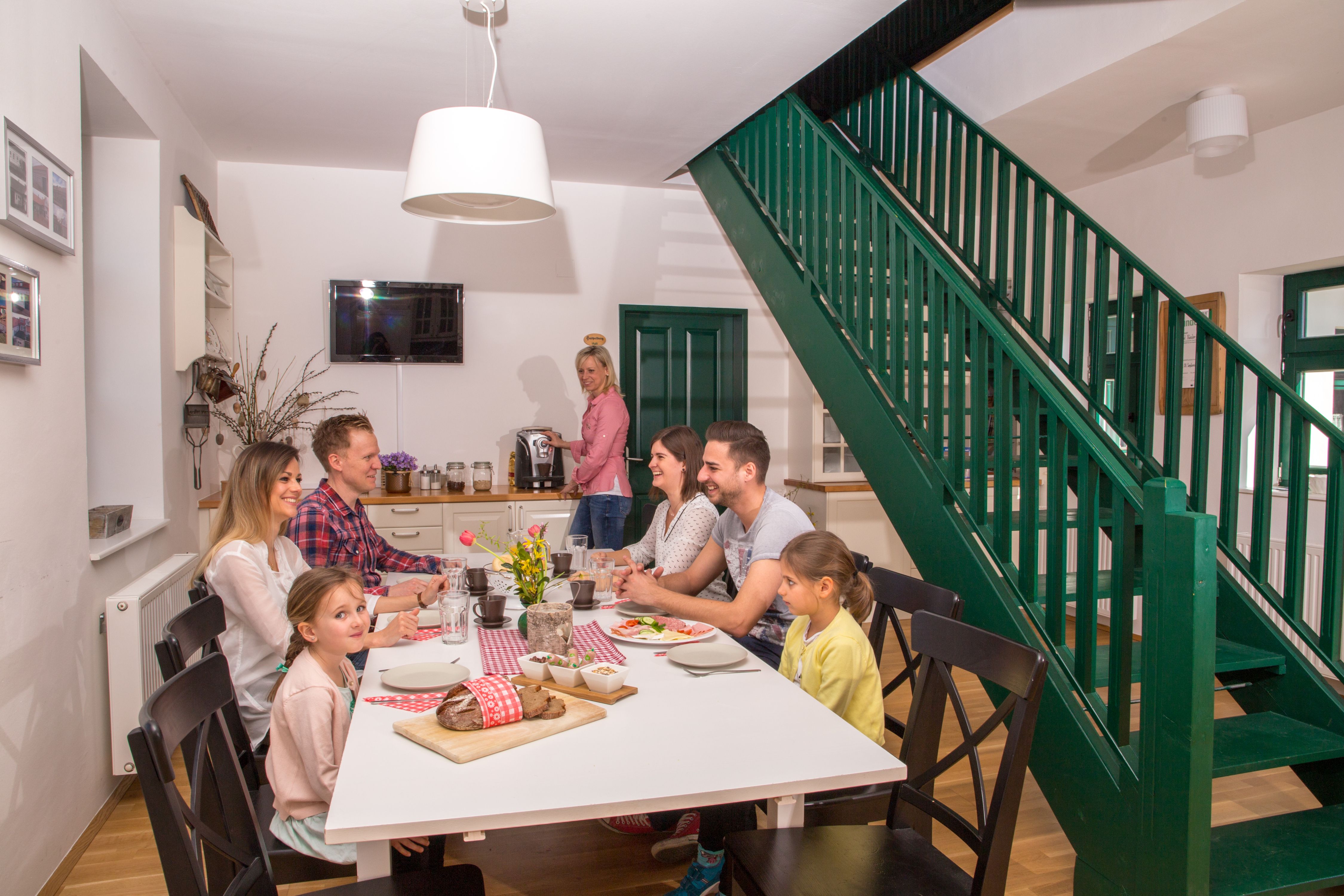 Rodina sedící u stolu v moderní jídelně se zeleným schodištěm.