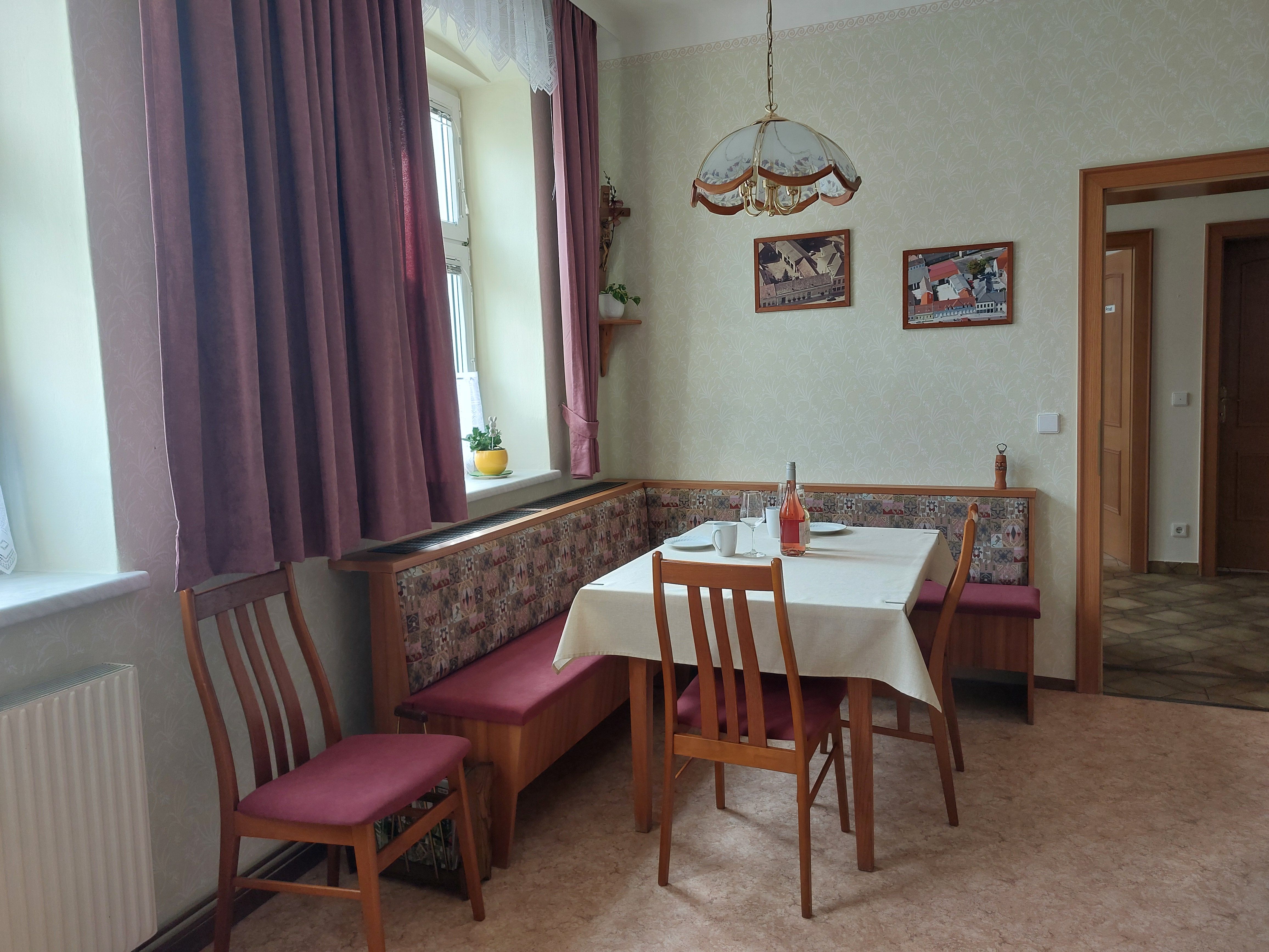 Útulná kuchyně s jídelním stolem, židlemi a rohovou lavicí. Okno se závěsy, obrázky na stěně.