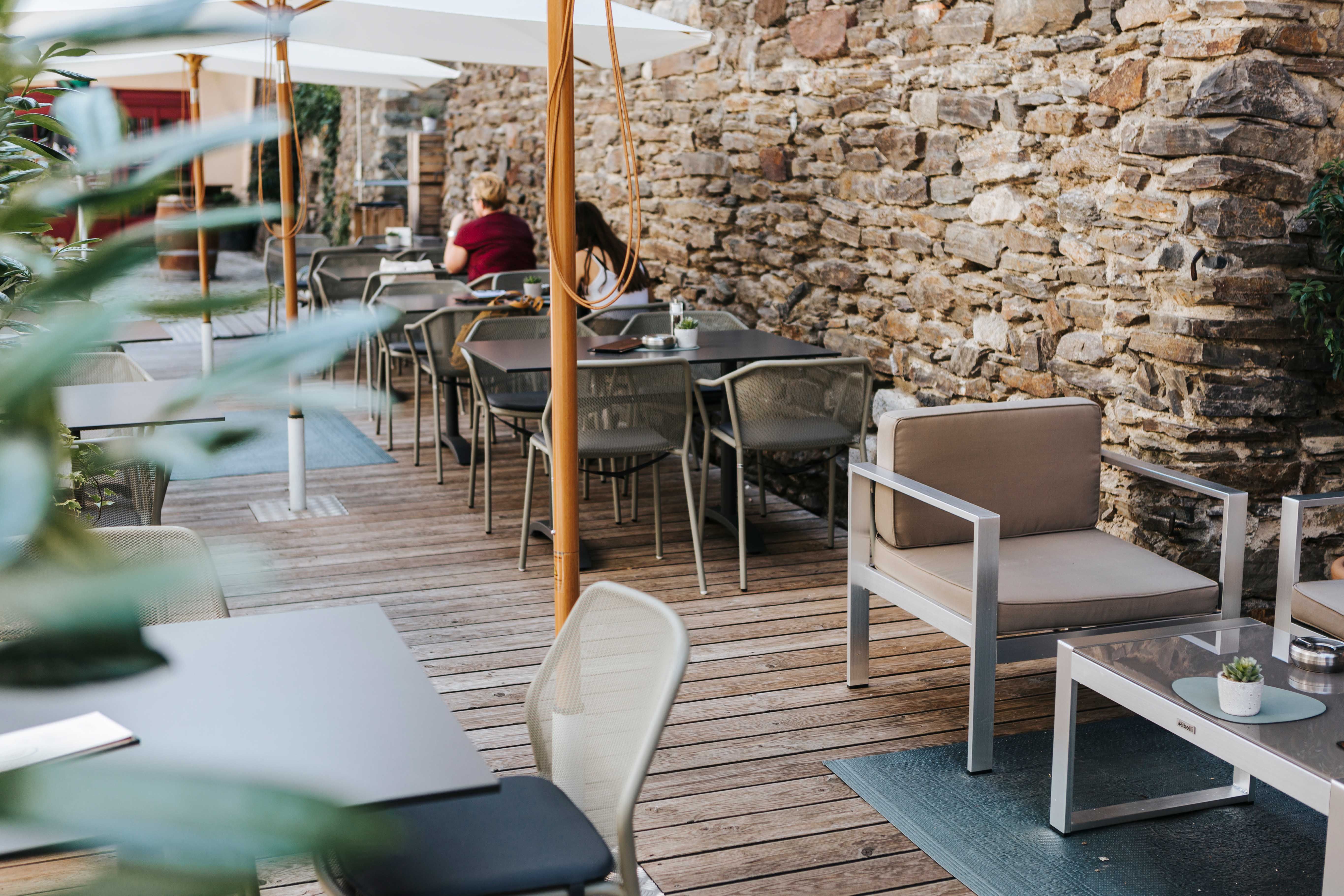 Útulný venkovní prostor kavárny s dřevěnou podlahou, moderním nábytkem a kamennou zdí.