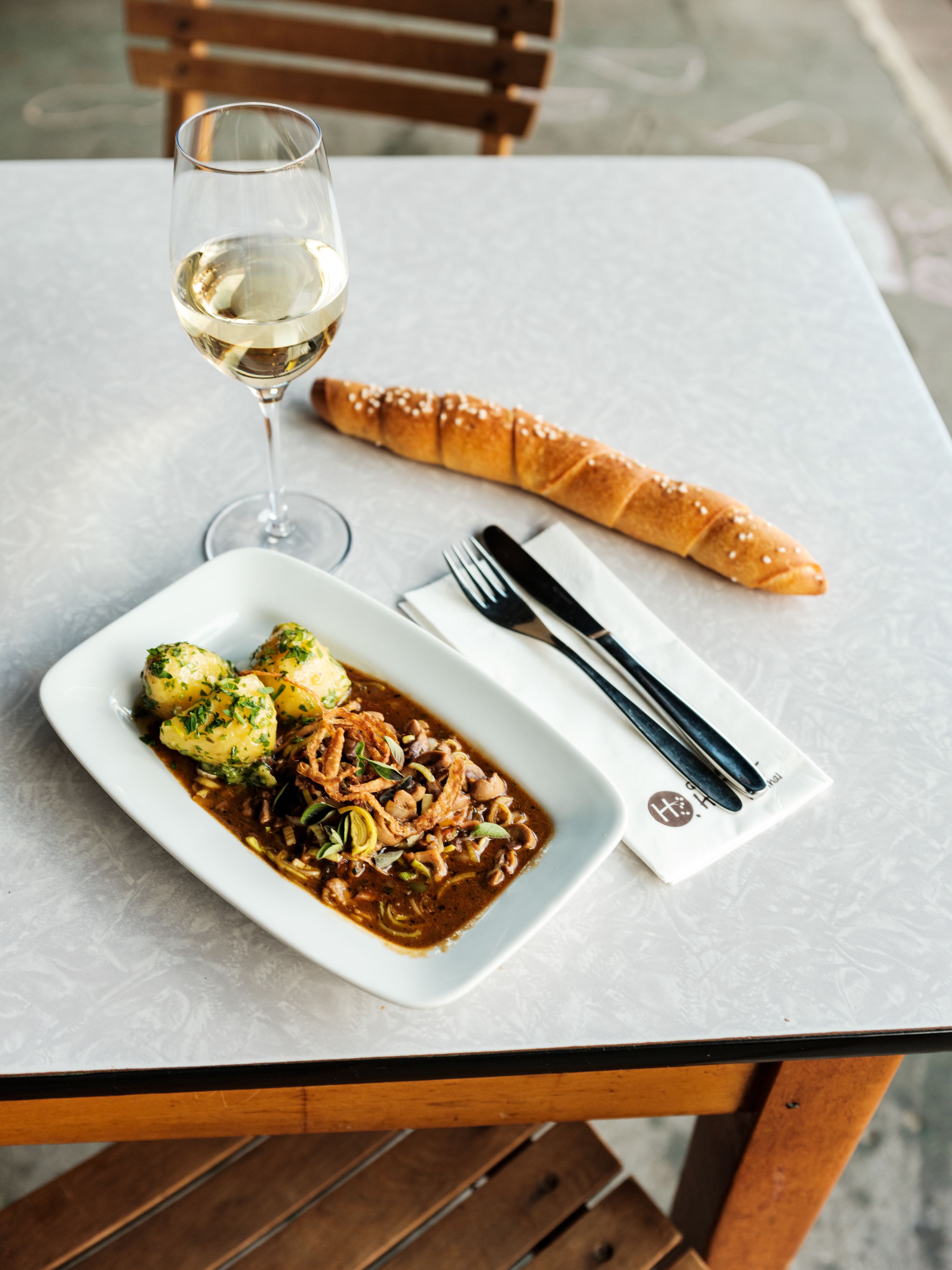 Stůl se sklenkou bílého vína, talíř s masovým pokrmem a knedlíky, dlouhá houska a příbor na ubrousku.