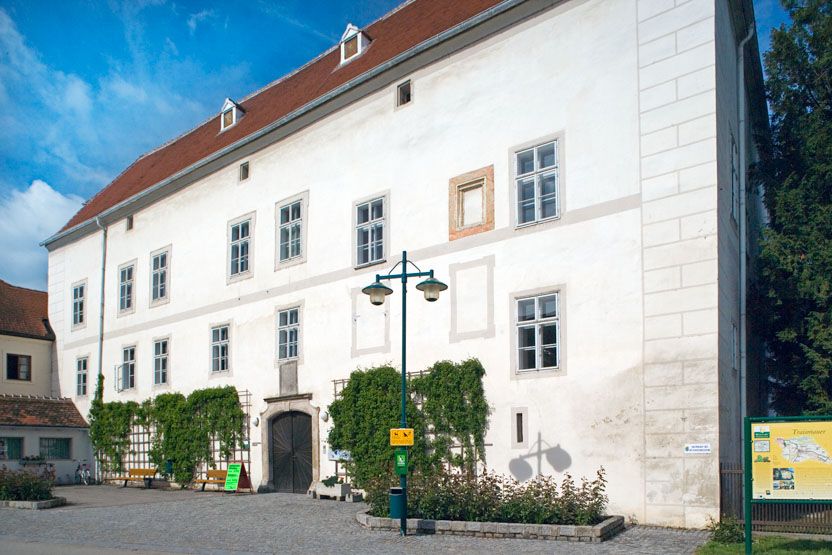 Zámek Traismauer s bílou fasádou a červenými střešními taškami, obklopený rostlinami a dlážděným předhradím.