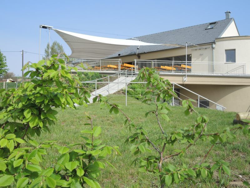 Vinárna s terasou a markýzou, obklopená zelení.