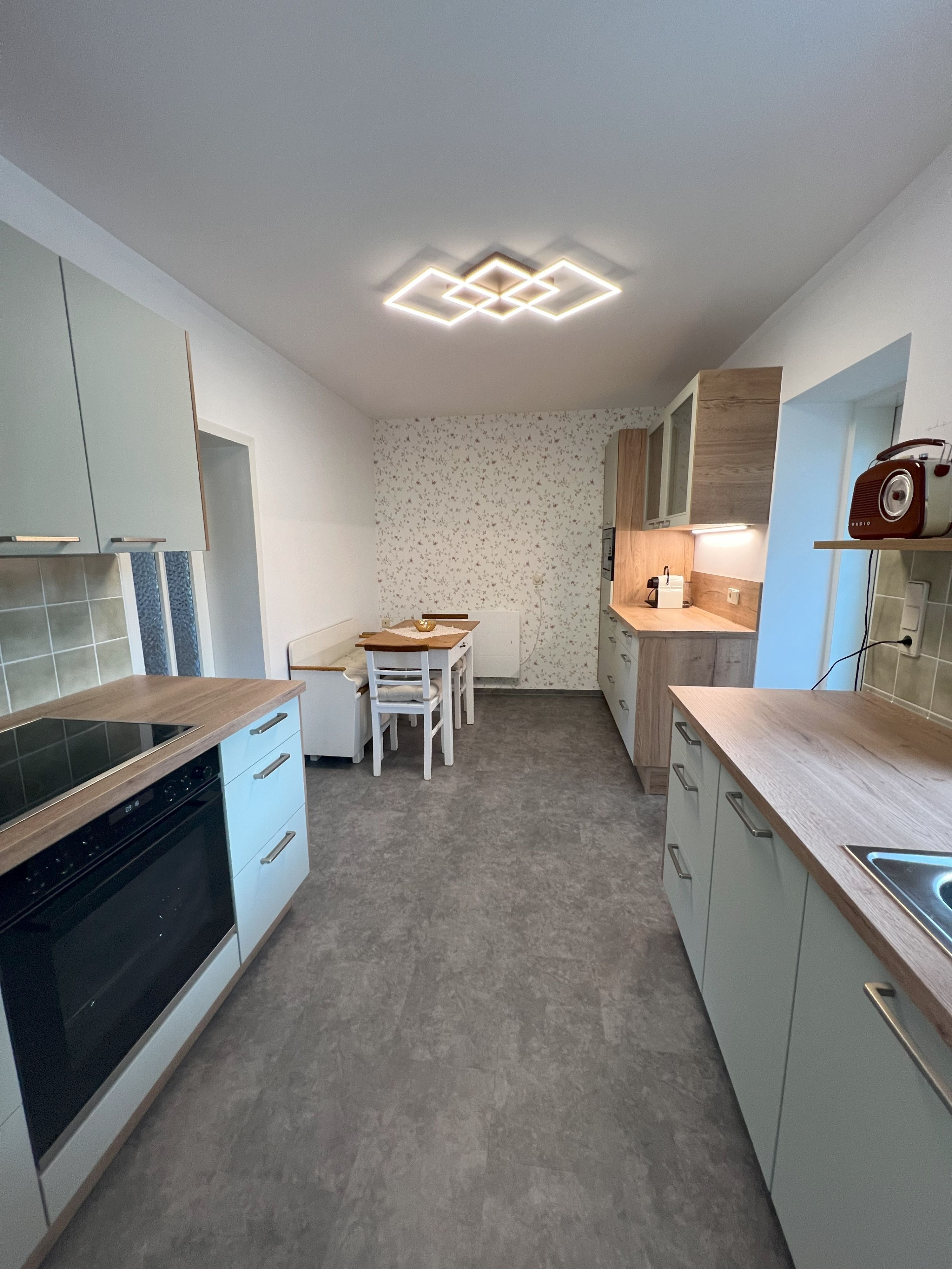 Moderní kuchyně s šedými skříňkami, dřevěnými akcenty a jídelním stolem. Geometrická lampa na stropě.