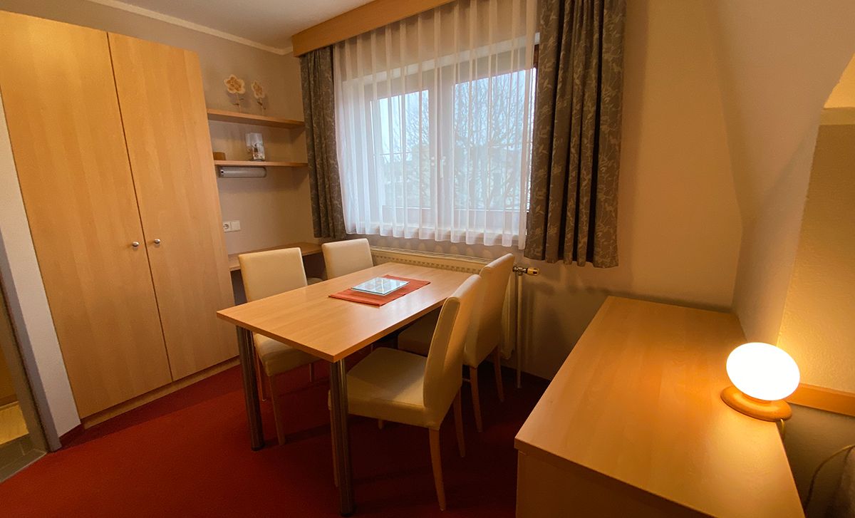 Útulný pokoj s dřevěným nábytkem, stolem, židlemi a oknem se závěsy.