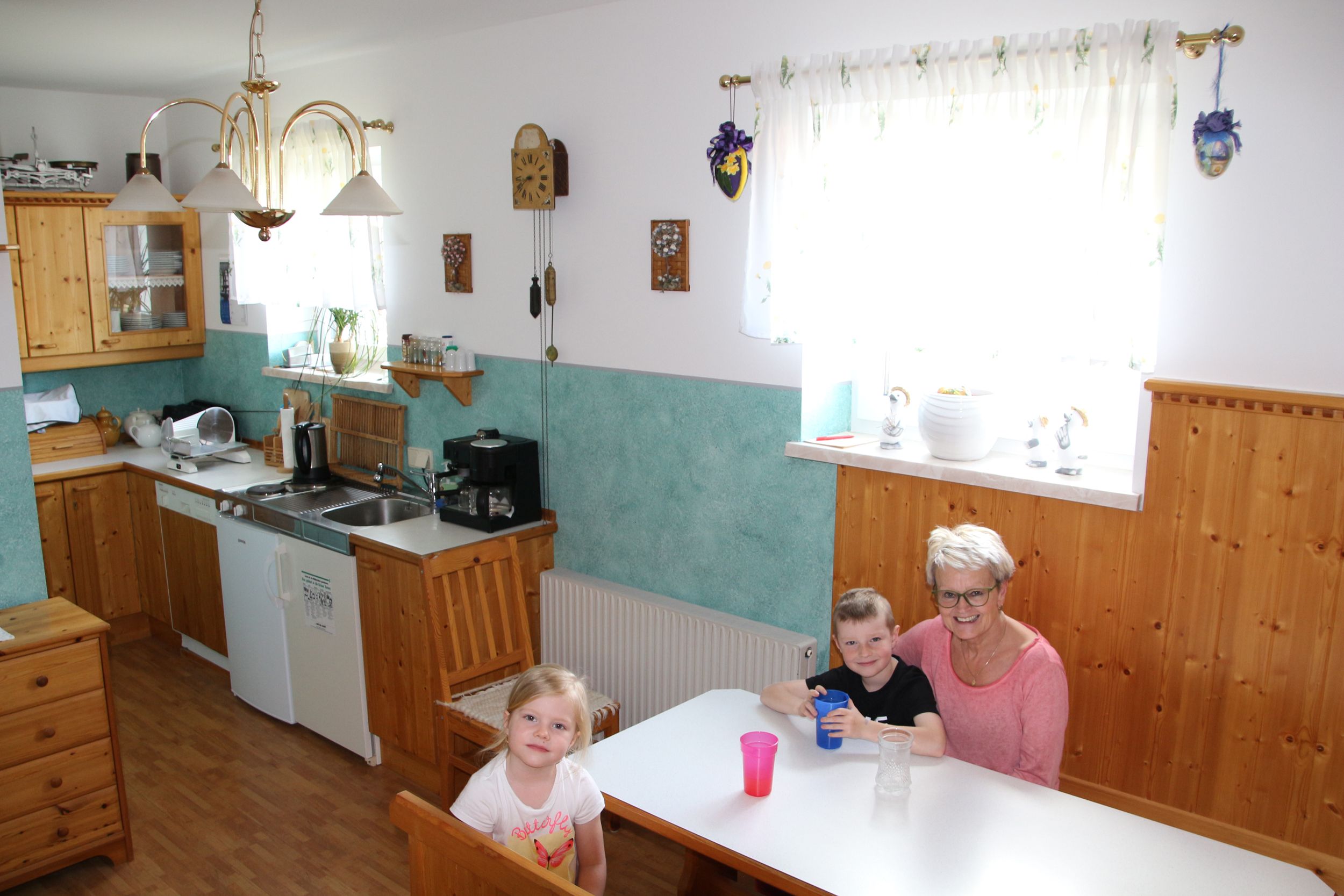 Snídaňová místnost s dřevěným nábytkem, kuchyní v pozadí a třemi osobami u stolu.