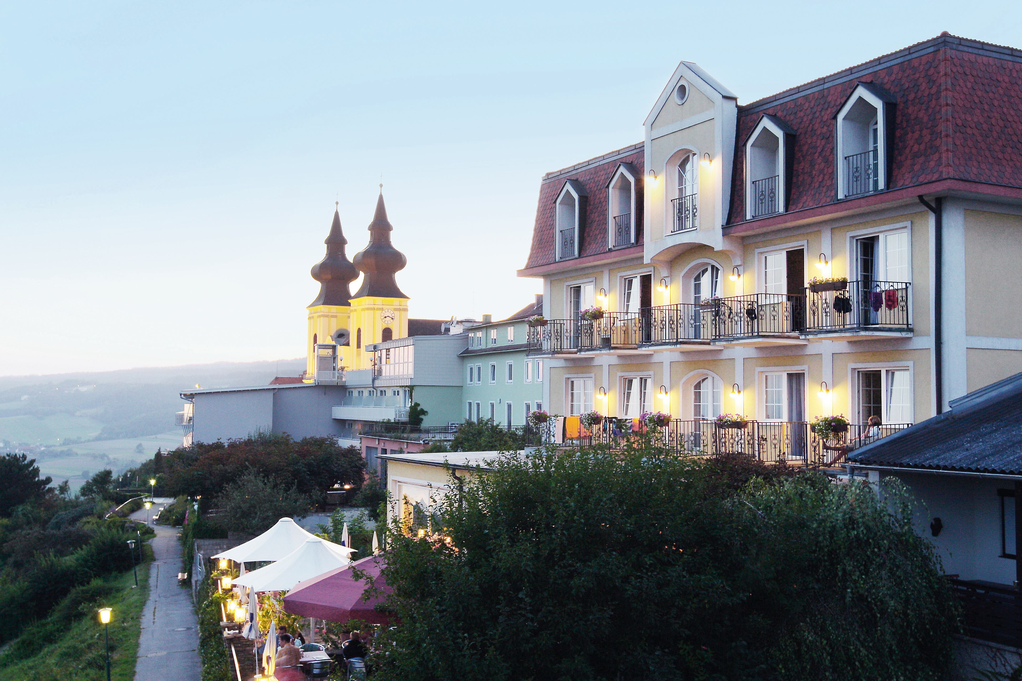 Hotel s balkony a kostelem v pozadí za soumraku.