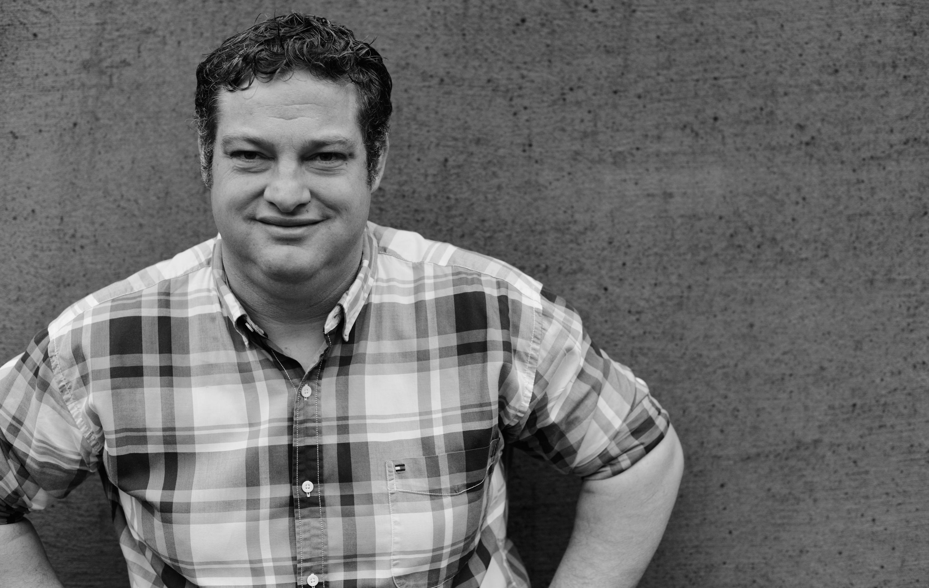 Černobílá fotografie muže v kostkované košili před betonovou zdí.
