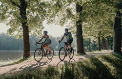 Zwei Personen, die man von vorne sieht, umgeben von Wald und Wasser, die mit ihren Rädern fahren