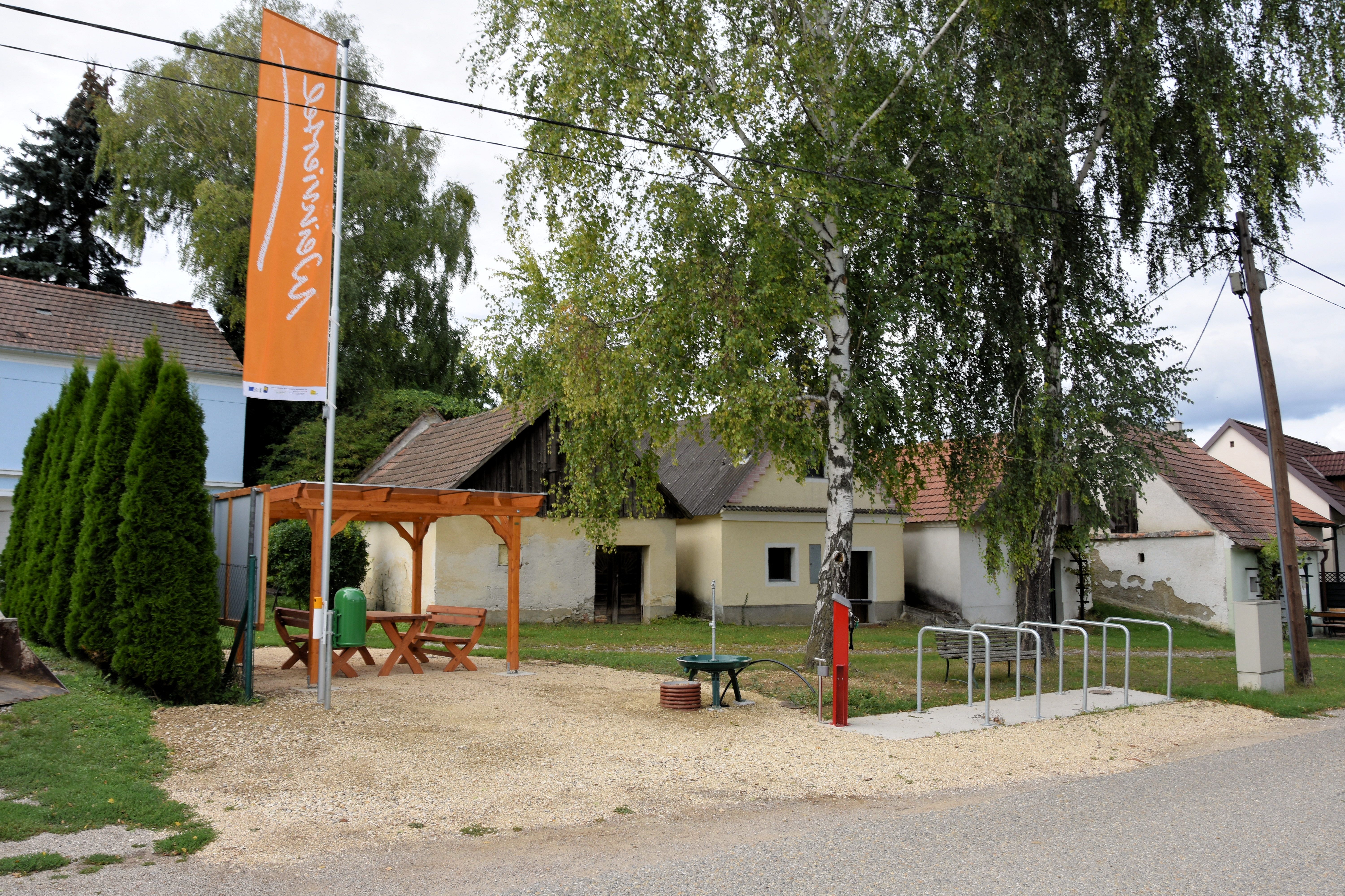 Odpočívadlo s dřevěným stolem a lavičkami pod altánem, obklopené stromy a starými budovami. K dispozici je také stojan na kola a informační tabule.