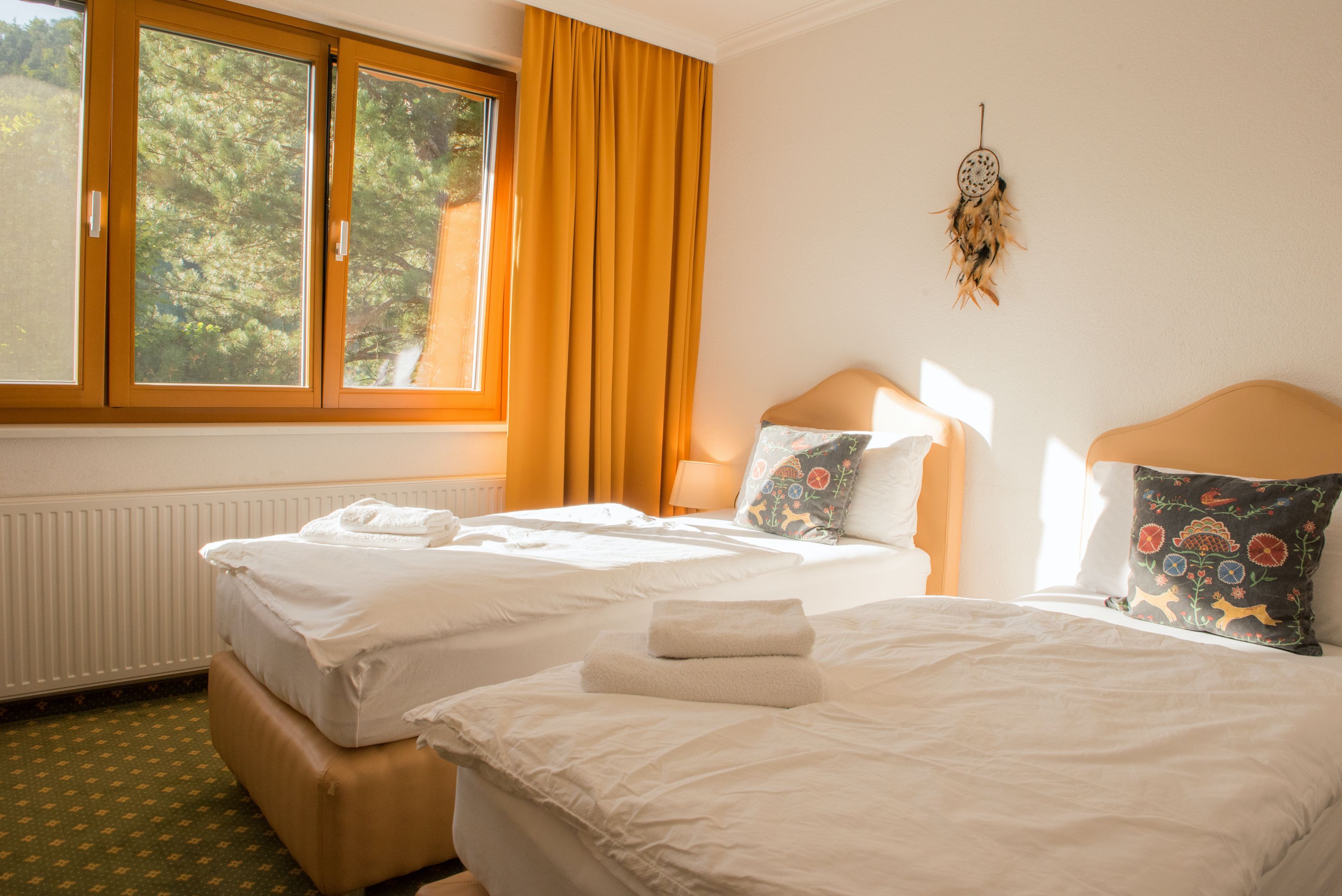 Světlý dvoulůžkový pokoj se dvěma samostatnými lůžky, žlutými závěsy a oknem s výhledem do zeleně.