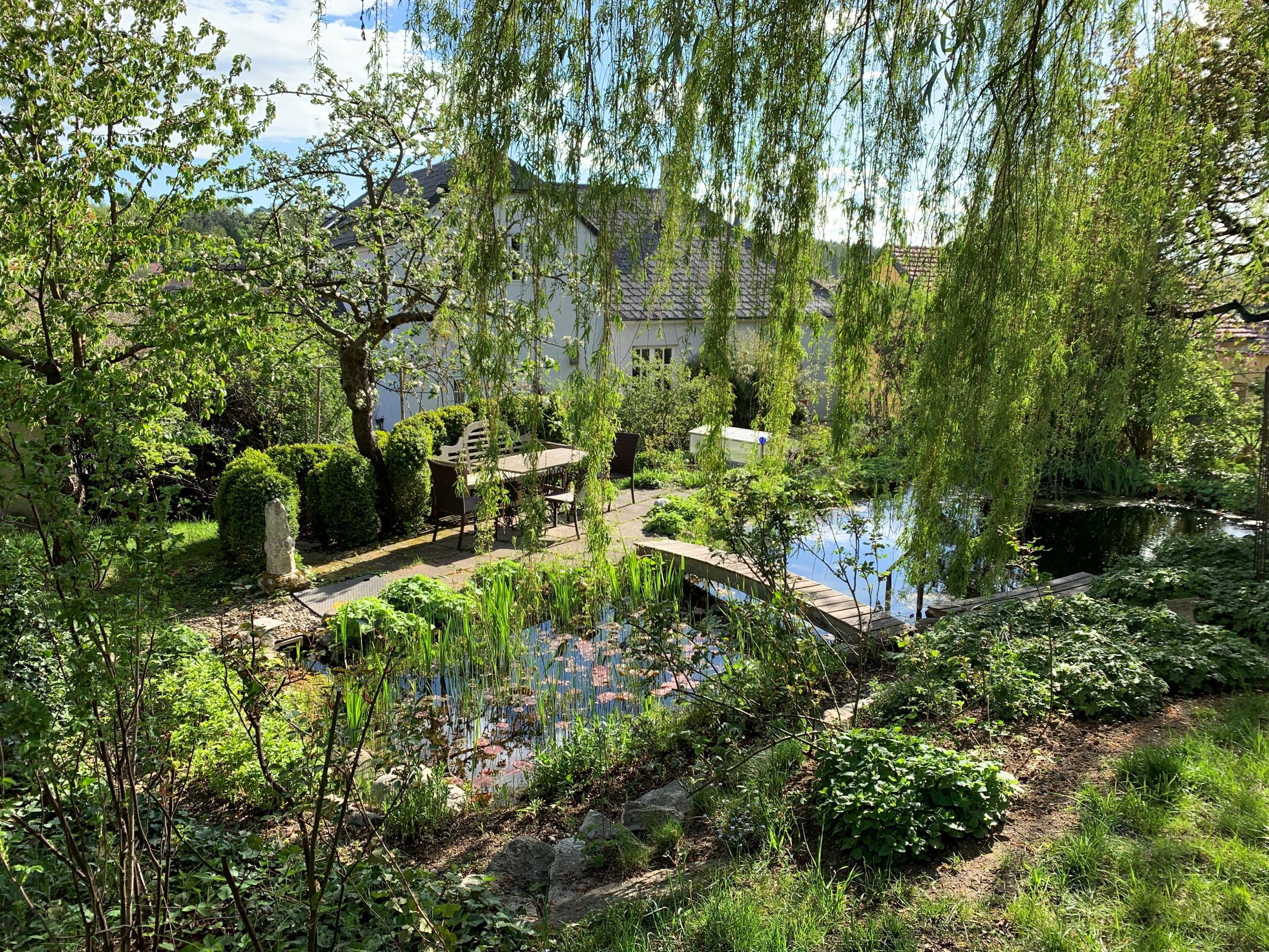 Idylická zahrada s jezírkem, stromy a stolem a židlemi pod širým nebem.