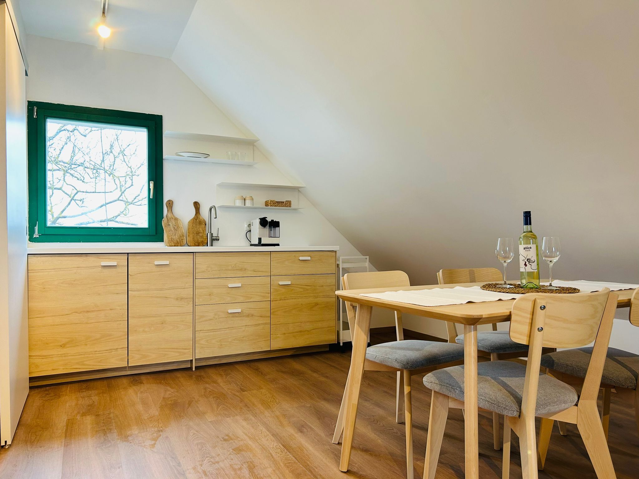 Moderní kuchyně s dřevěnými skříňkami, jídelní stůl a židle, okno se zeleným rámem.