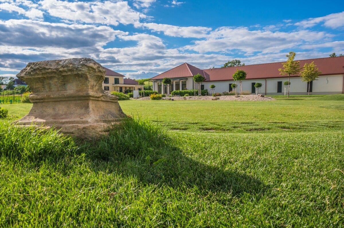 Vinařství s trávníkem a antickým kamenem v popředí.