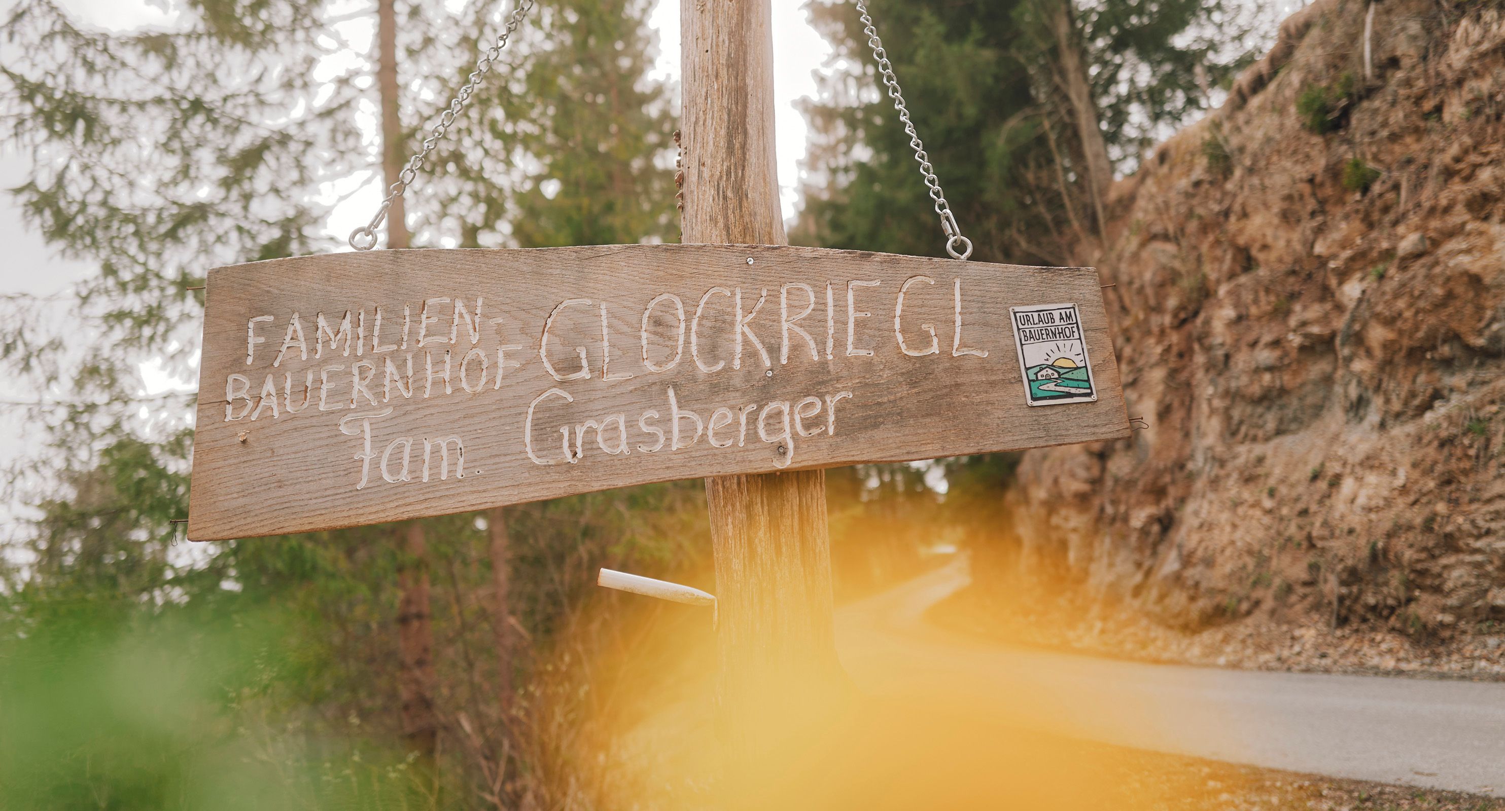 Dřevěná cedule s nápisem "Familienbauernhof Glockriegl Fam. Grasberger" na lesní cestě.