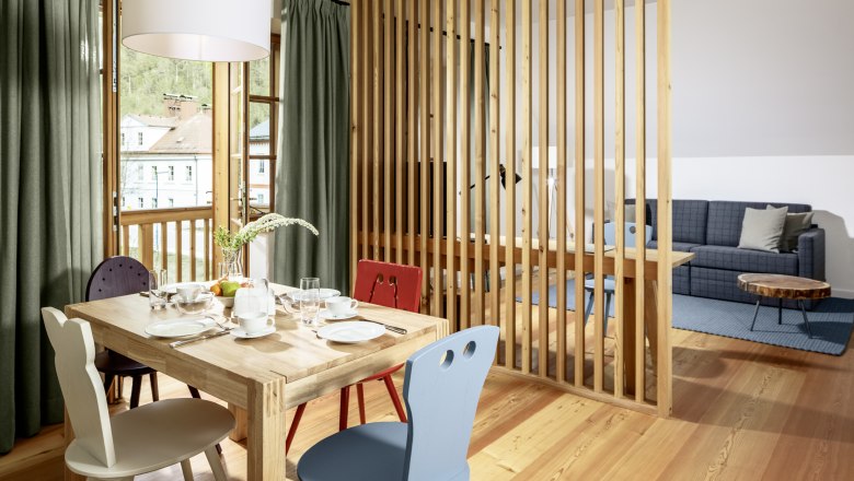 Světlý hotelový pokoj s dřevěnou podlahou, jídelním stolem a židlemi, oddělený dřevěnou lamelovou stěnou, s výhledem na pohovku a konferenční stolek.