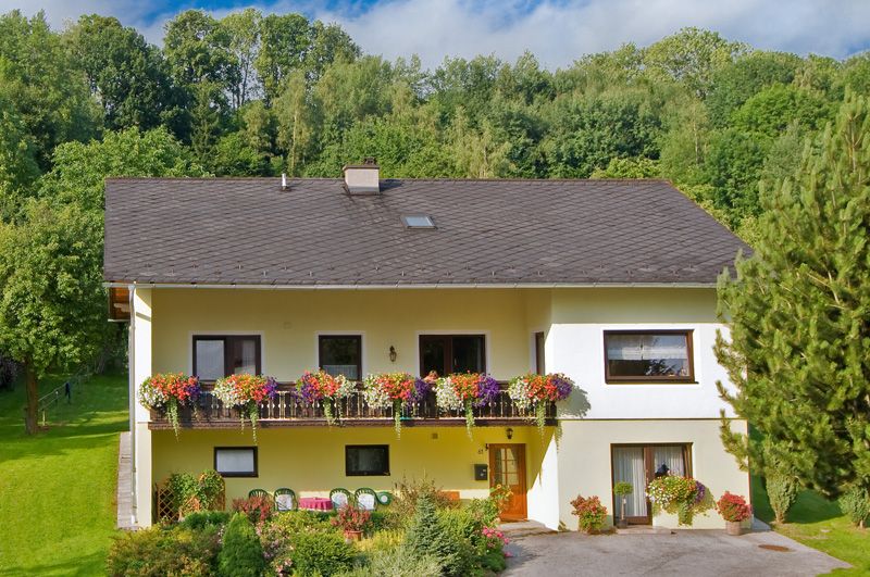 Dvoupatrový žlutý dům s balkonem a květinami, obklopený stromy a zahradou.