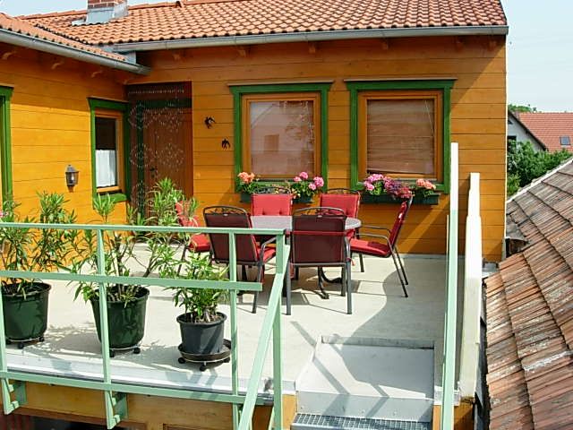 Balkon se stolem, židlemi a rostlinami, obklopený domem s oranžovou fasádou a zelenými okenními rámy.