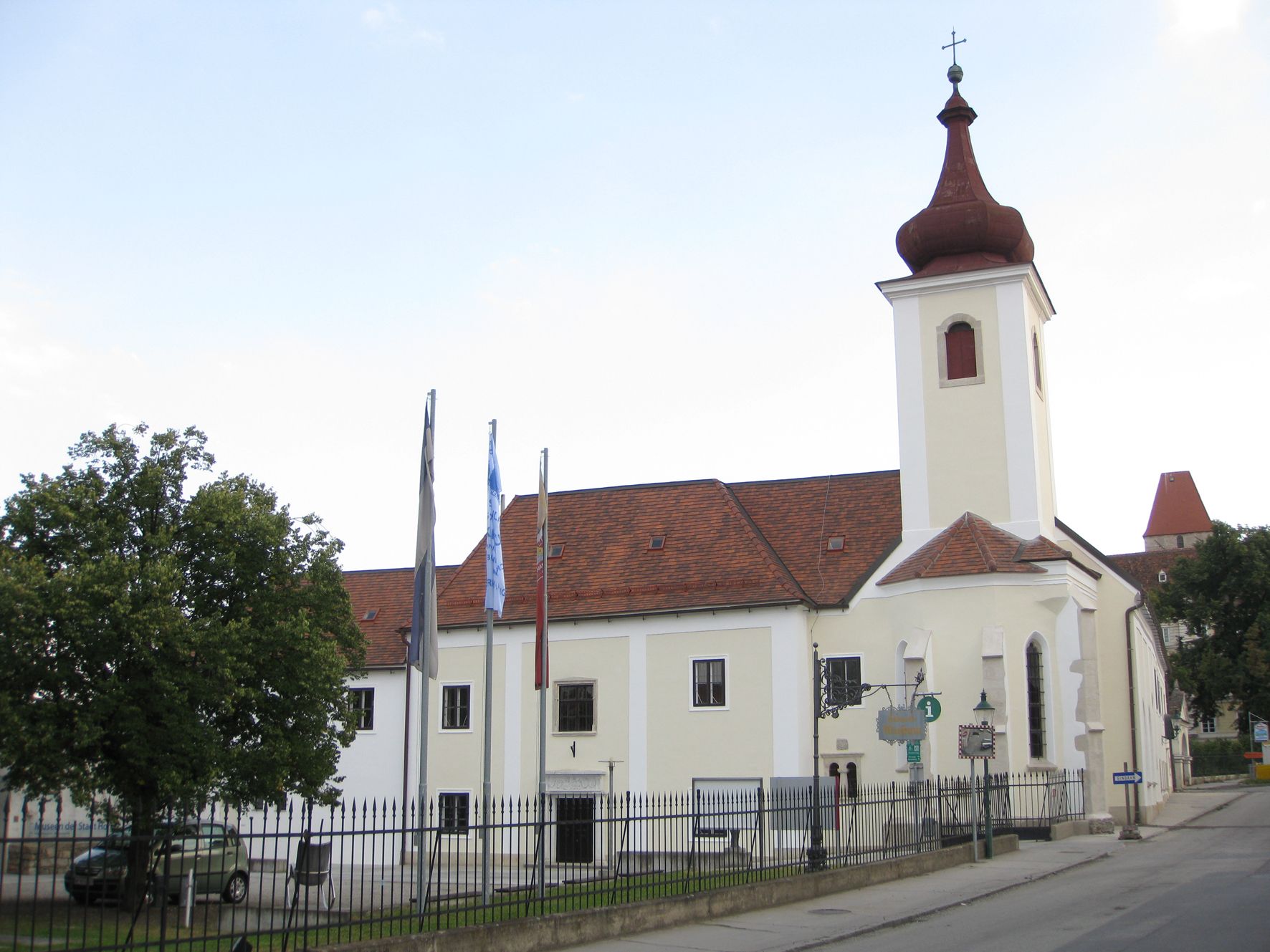 Budova s kostelní věží a červenými střešními taškami, obklopená plotem a stromy.