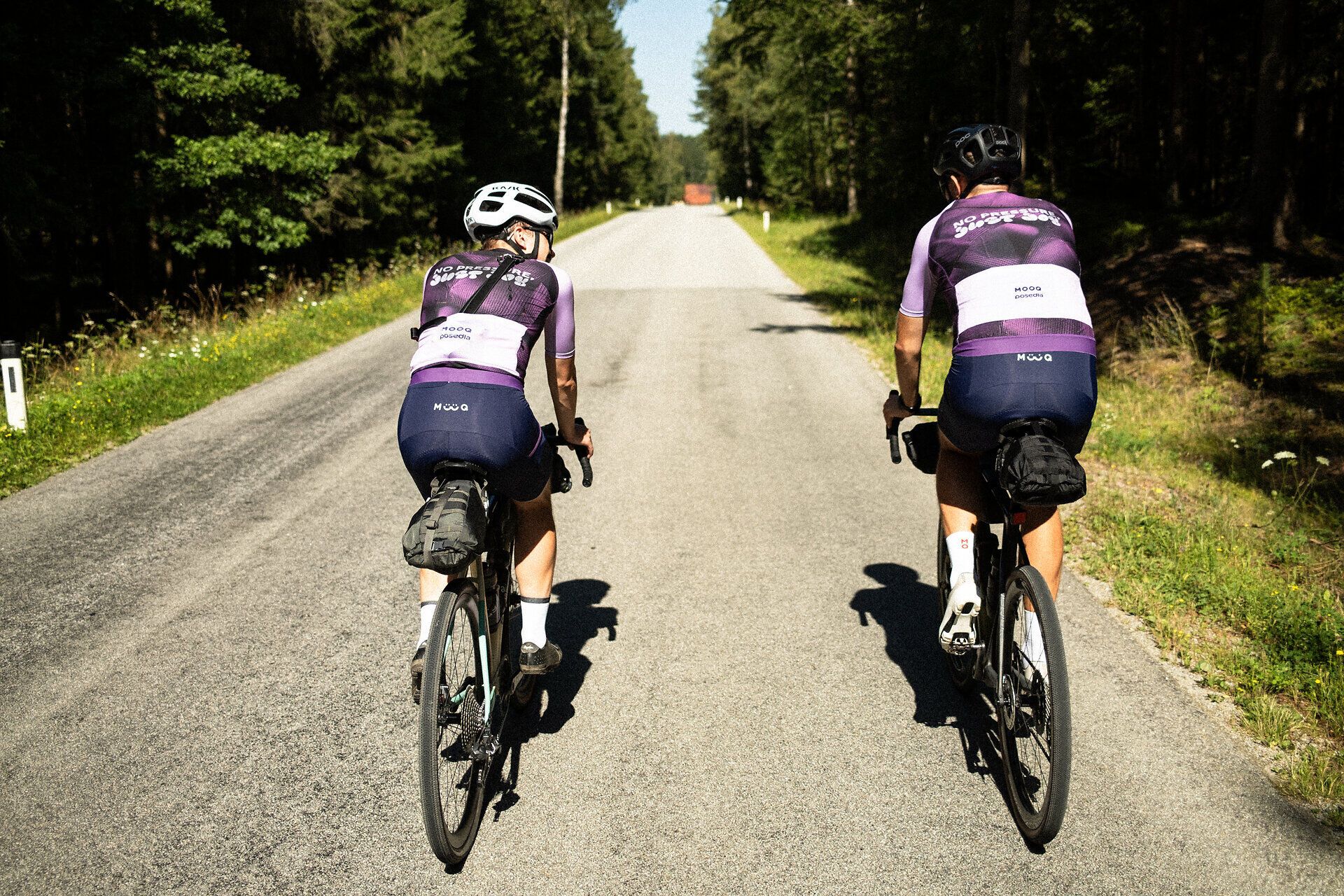 Dva cyklisté si užívají čerstvého vzduchu a klidné atmosféry na malebné venkovské silnici obklopené bujnou zelení a vzrostlými stromy. Zvlněné kopce v pozadí vybízejí k objevování krás přírody a prožívání svobody jízdy na kole.