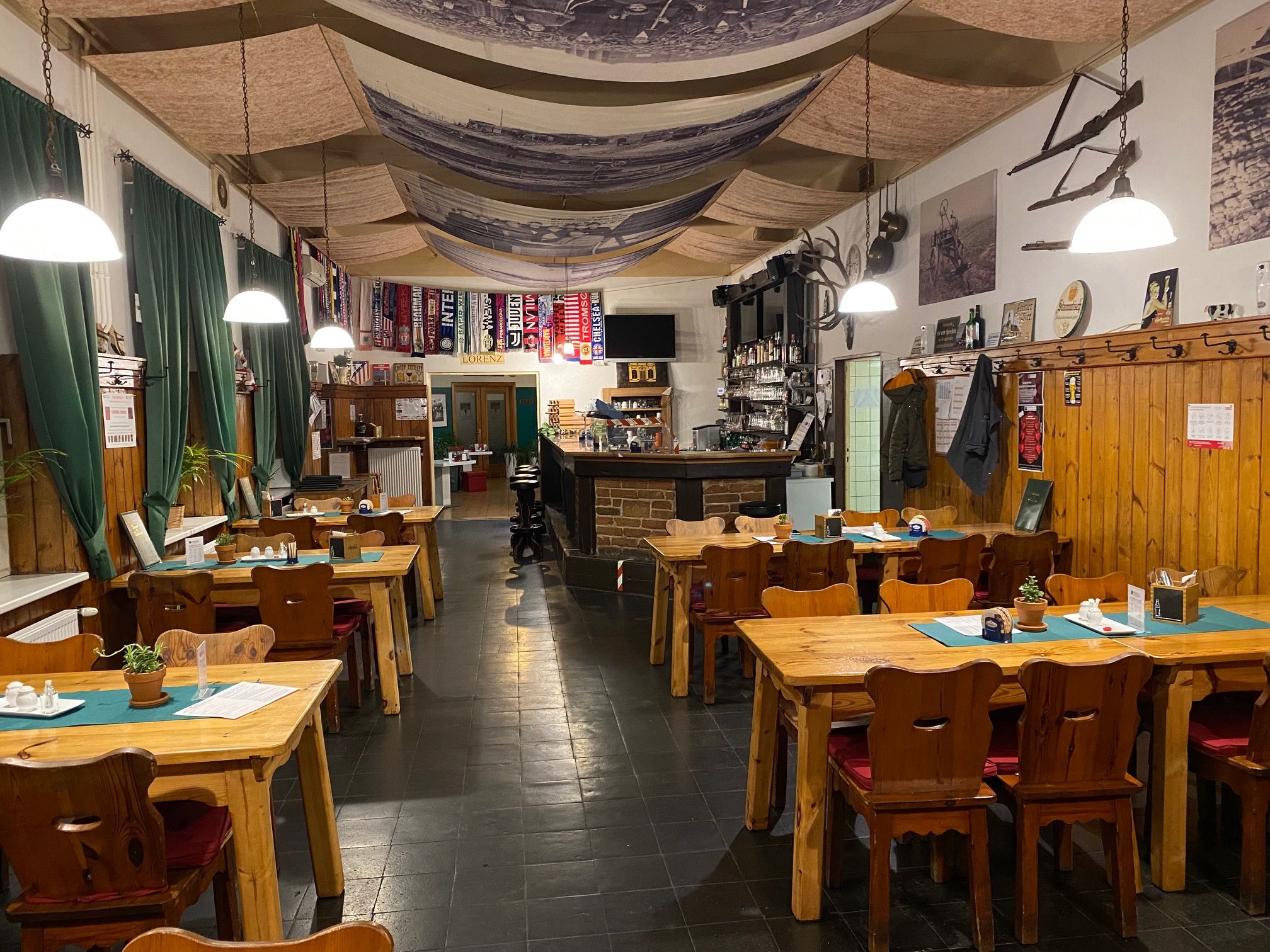 Útulná restaurace s dřevěnými stoly, židlemi a barem, vyzdobená fotbalovými šálami a rustikálními prvky.