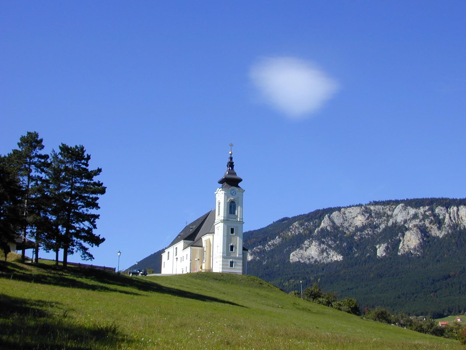 Kostel na kopci s modrou oblohou a horami v pozadí.