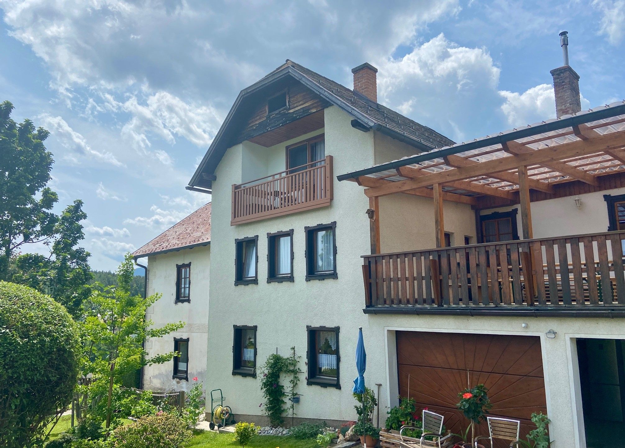 Dvoupatrový dům s balkonem a terasou, obklopený zahradou a stromy, pod zamračenou oblohou.