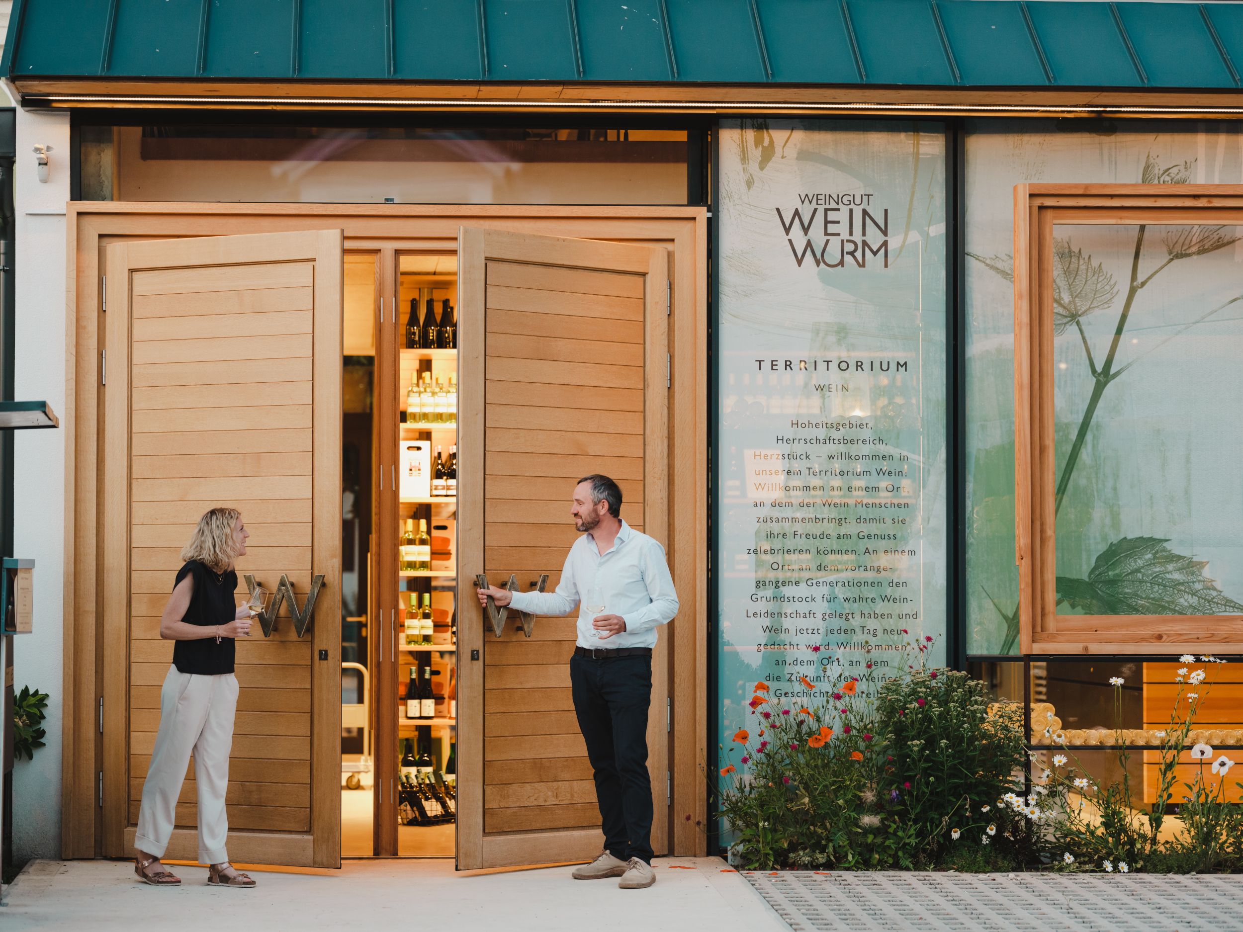 Dva lidé stojí před vinařstvím s otevřenými dřevěnými dveřmi, v nichž jsou vidět lahve.