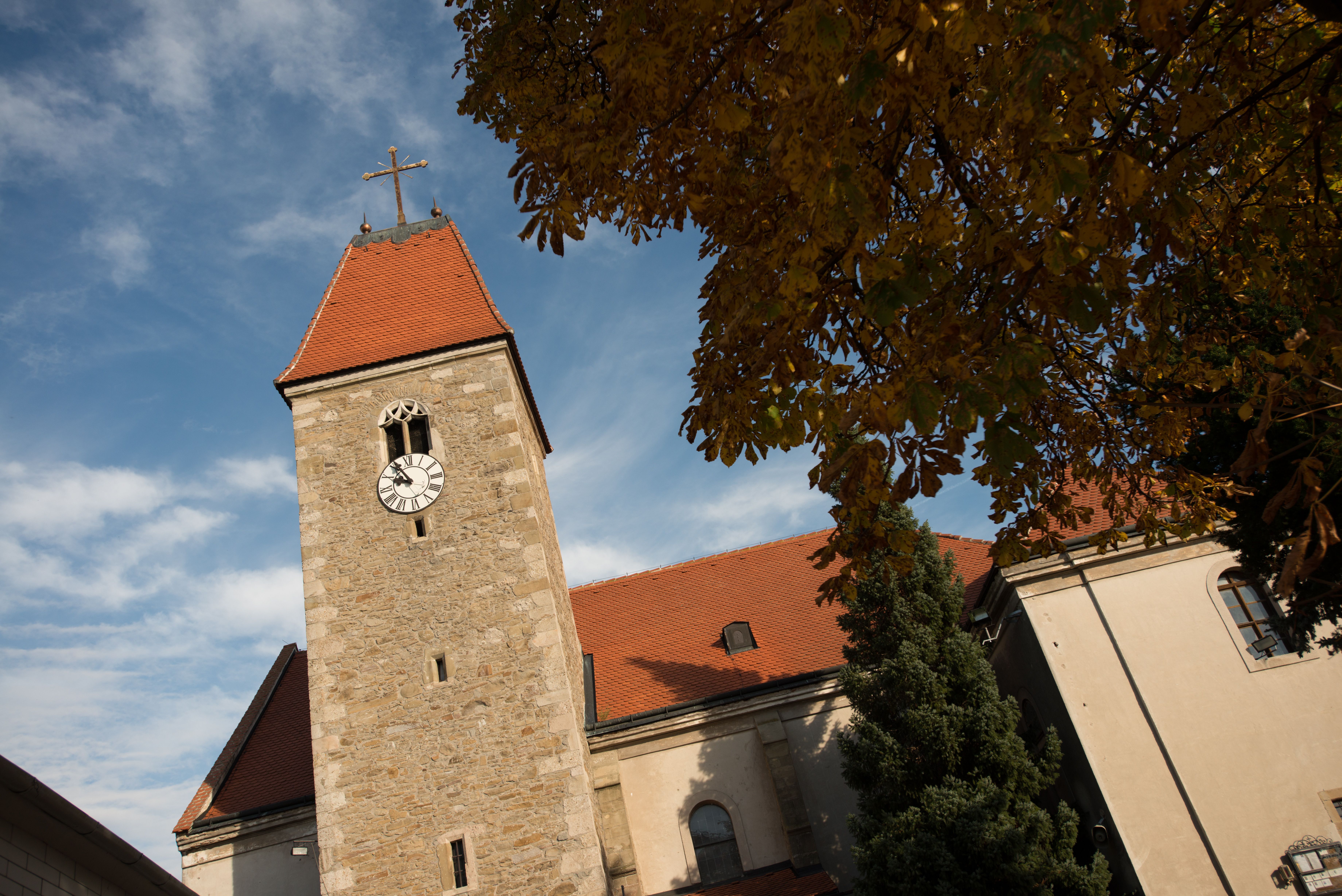 Věž kostela ve Weißenkirchenu s hodinami a červenou střechou, obklopená podzimním listím a modrou oblohou.