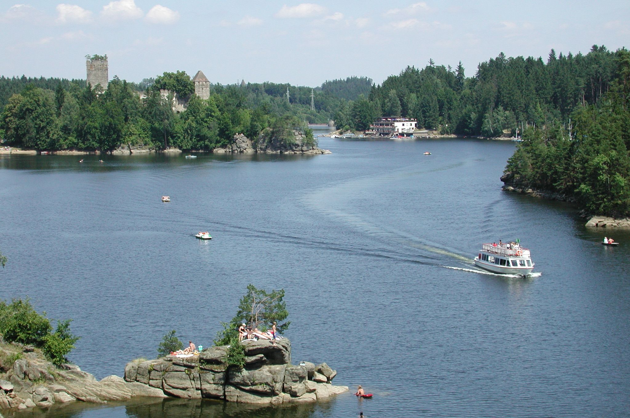 Jezero s loděmi, hrad v pozadí a lidé na skalách.