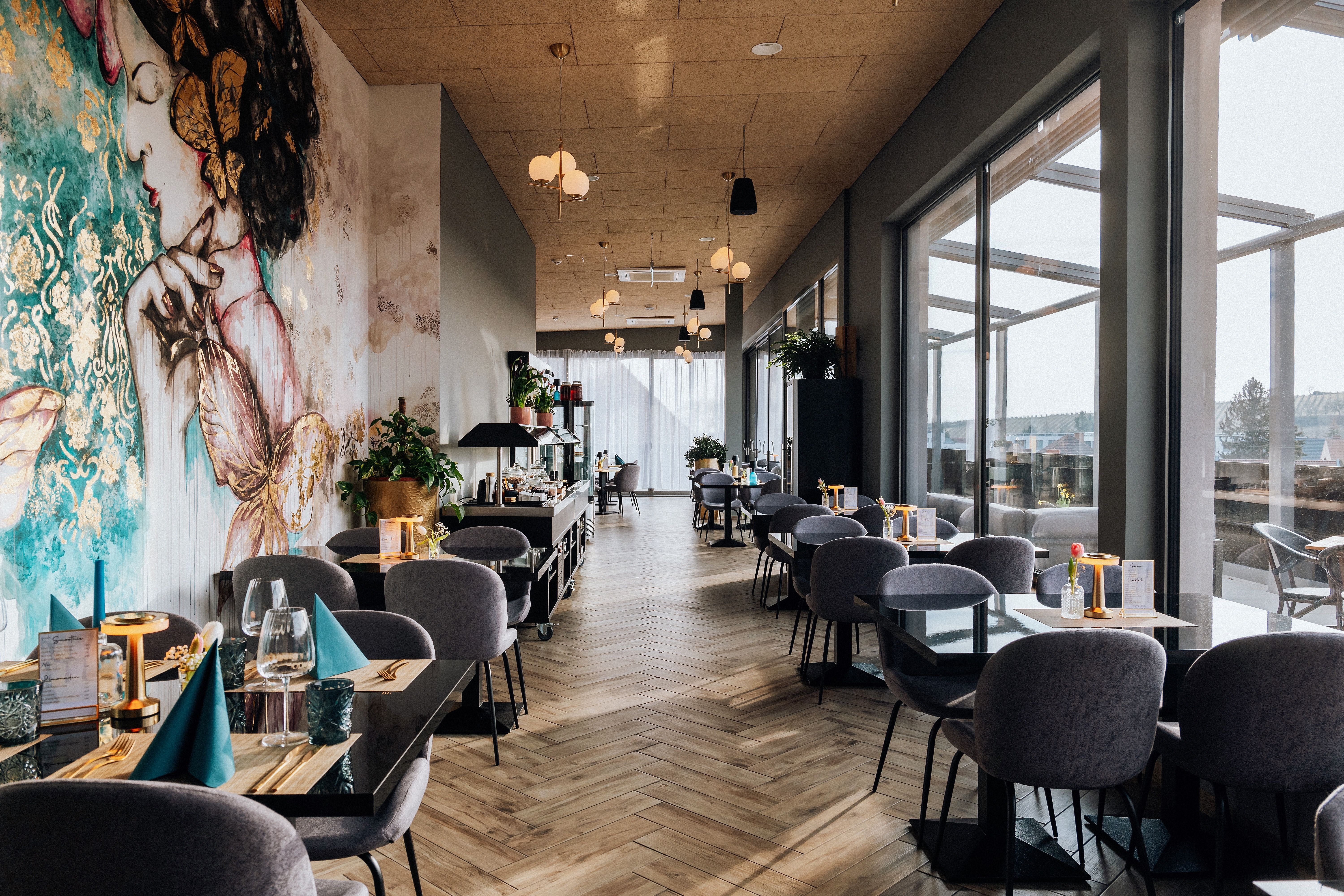 Pohled do interiéru stylové restaurace s velkými okny a uměleckými nástěnnými malbami.