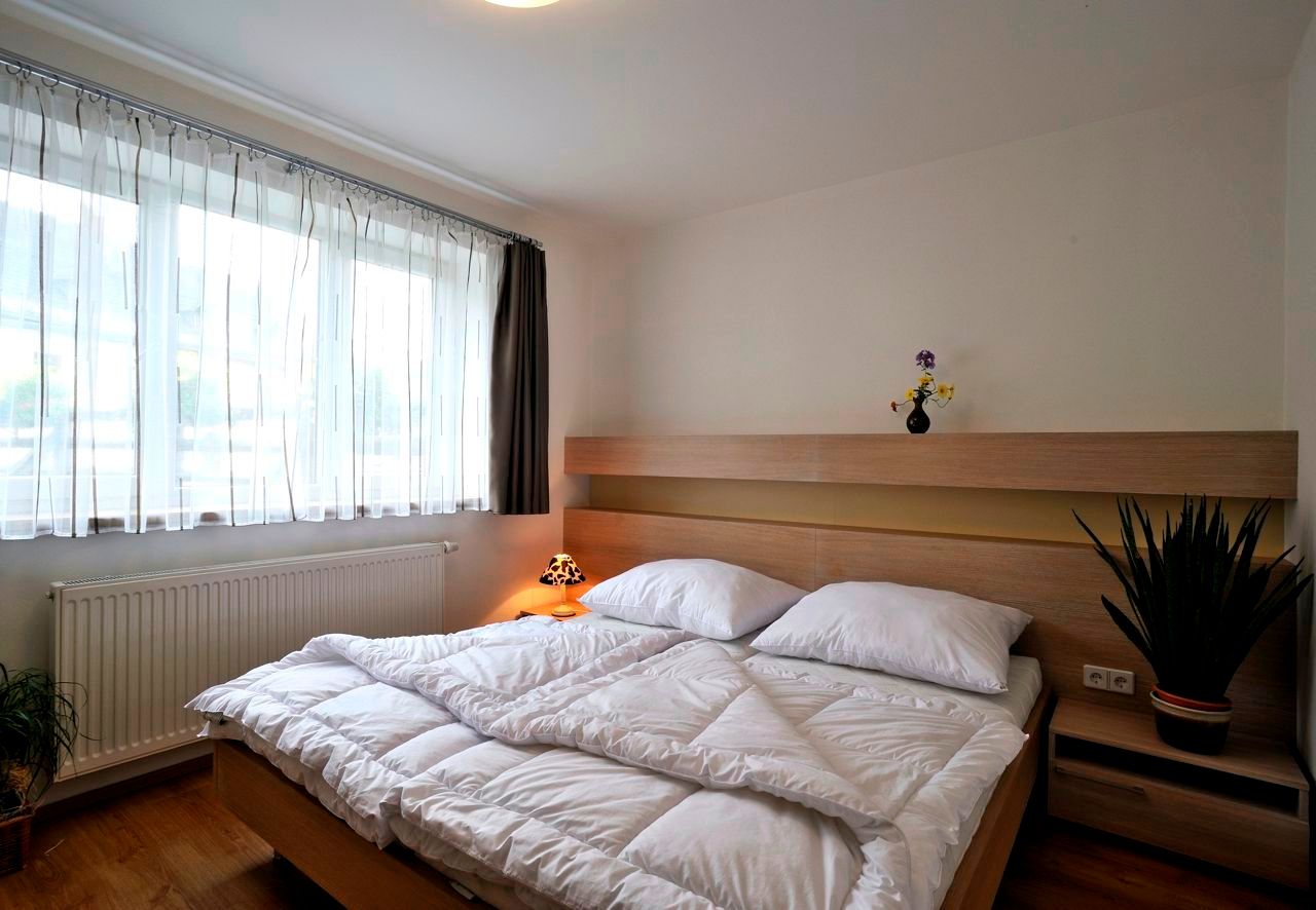Moderní ložnice s manželskou postelí, světlým dřevěným nábytkem a velkými okny s bílými závěsy.