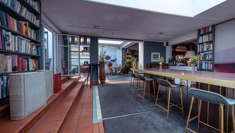 Pohled do interiéru moderního hotelového pokoje s policemi na knihy, vysokými stoly a barovými židlemi.