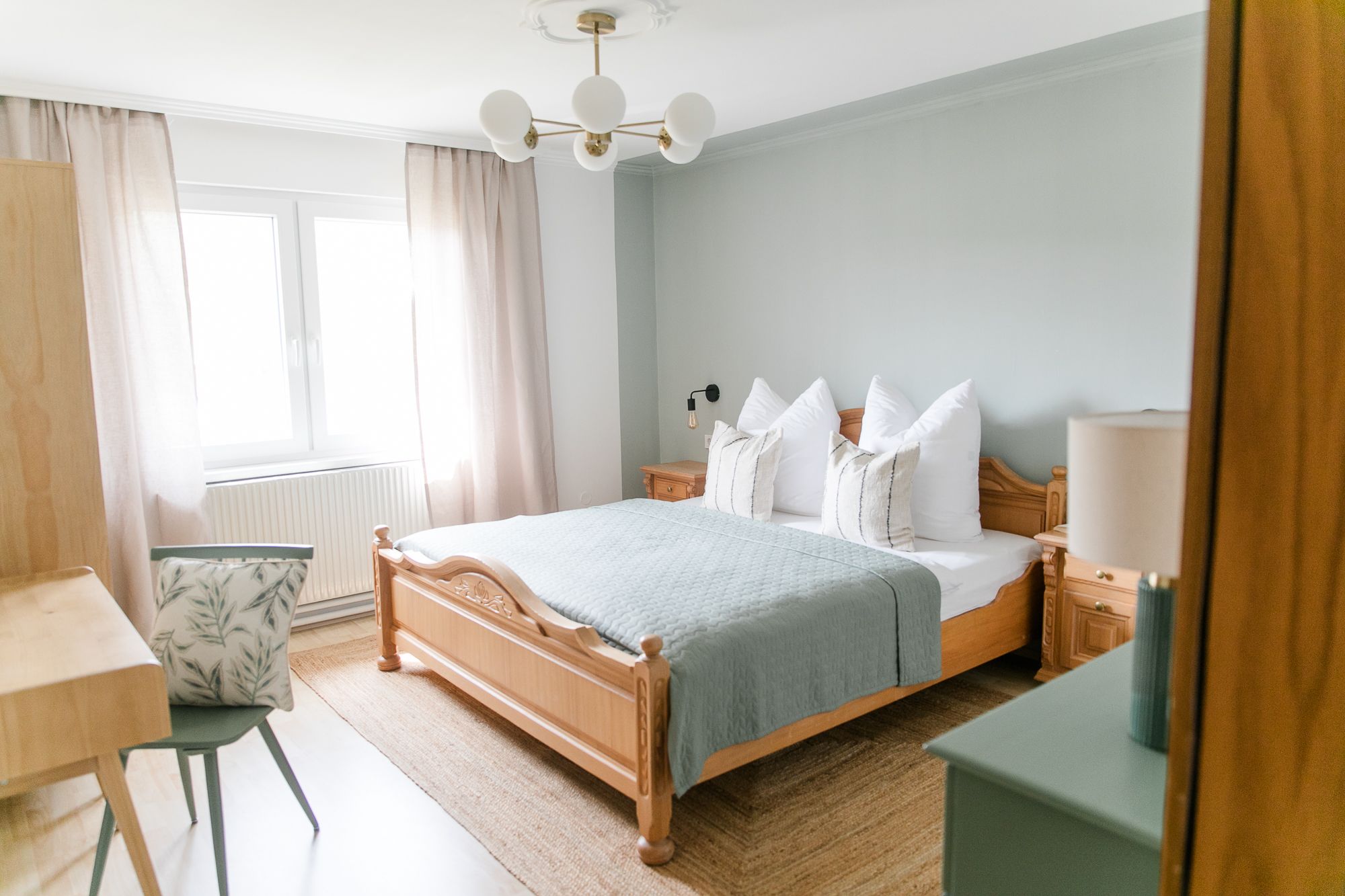 Útulná ložnice s dřevěnou postelí, modrou peřinou, nočními stolky a psacím stolem se židlí. Vidět jsou světlé závěsy a okno.