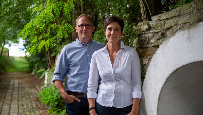 Muž a žena stojí vedle sebe na dlážděné cestě pod širým nebem, obklopeni stromy a kamennou zdí.