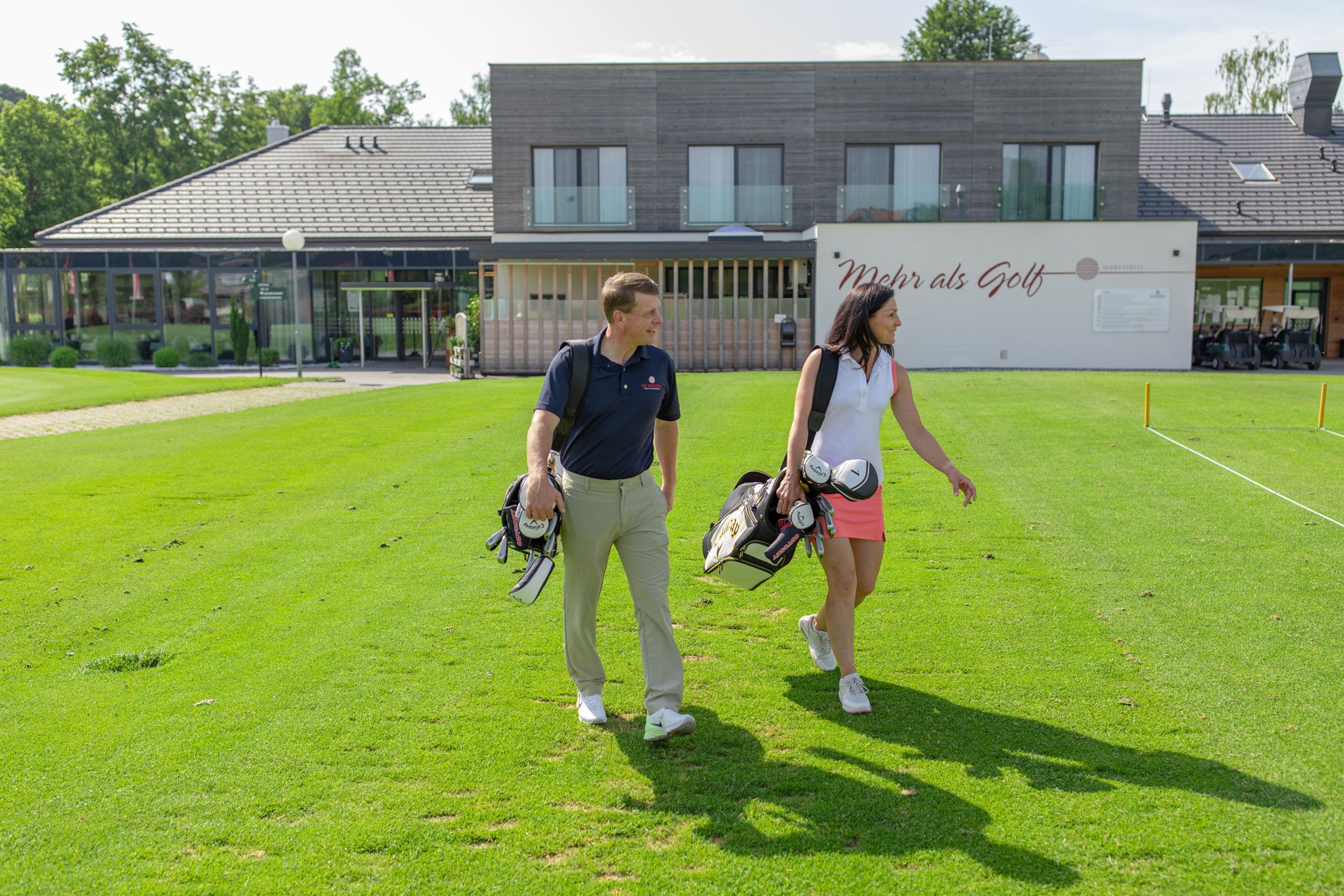 Dva lidé s golfovými bagy před moderní klubovnou.