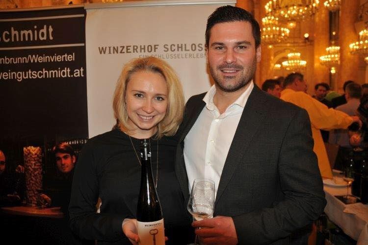 Pár drží láhev vína a sklenici vína před transparentem s nápisem "Winzerhof Schlosser".