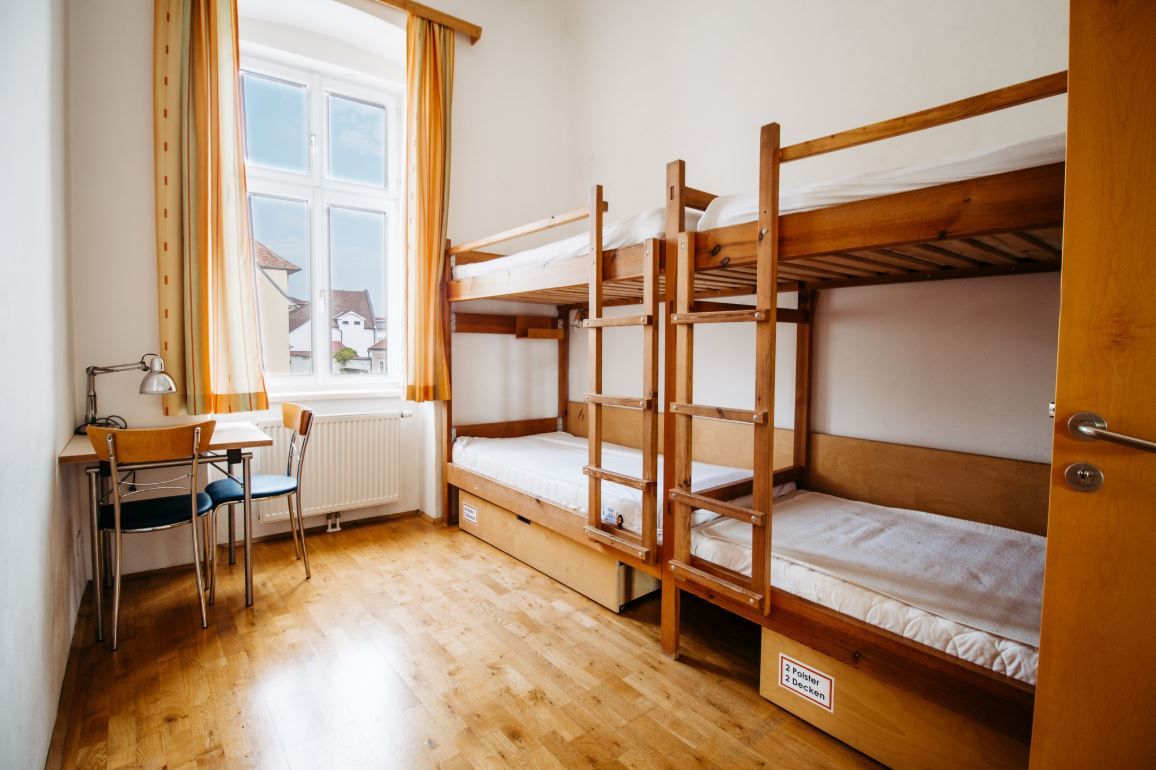 Jednoduchý pokoj s palandou, stolem a židlí, světlé závěsy u okna.