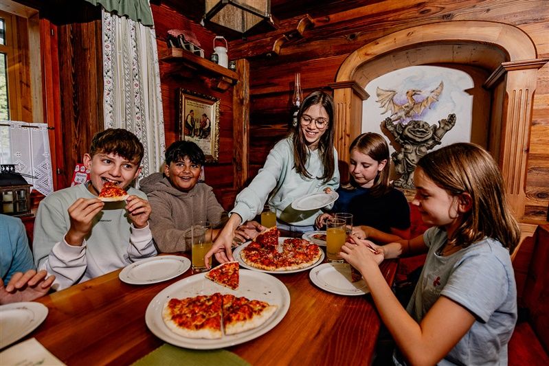 Děti jedí pizzu v rustikální místnosti s dřevěnými stěnami.