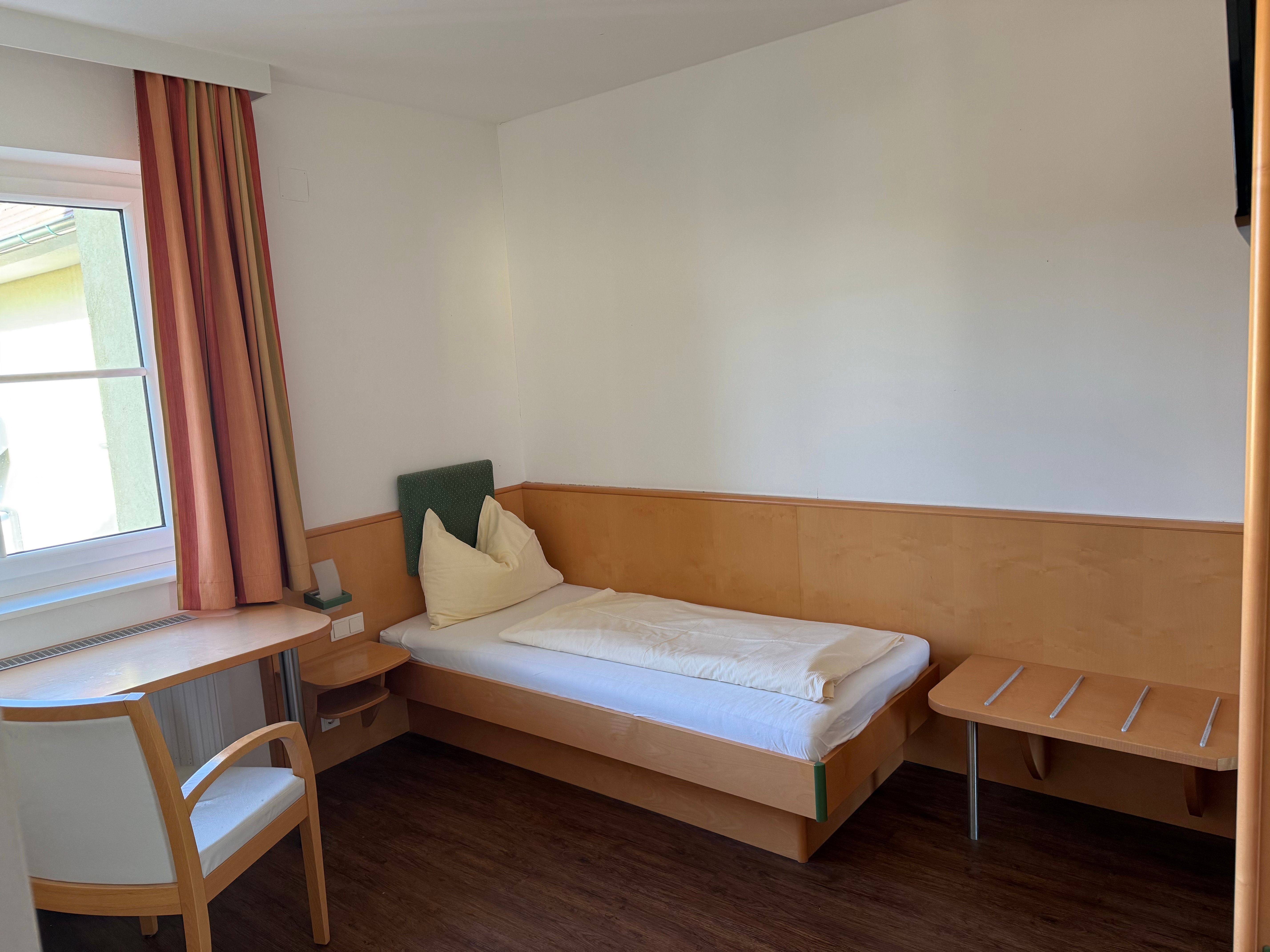 Jednoduchý hotelový pokoj s jednou postelí, stolem, židlí a oknem se závěsy.