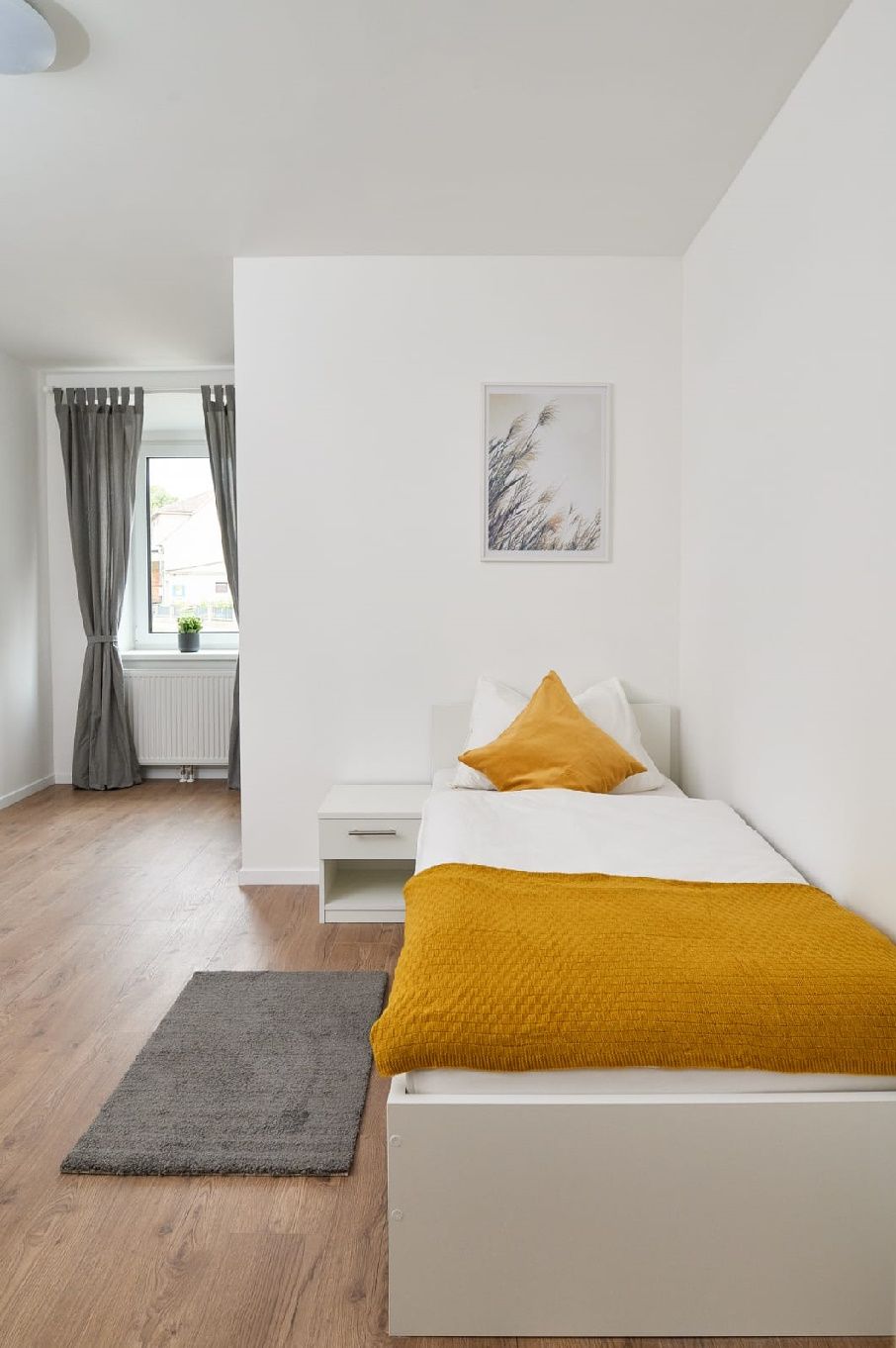 Jednolůžkový pokoj s postelí, nočním stolkem a oknem se závěsy.